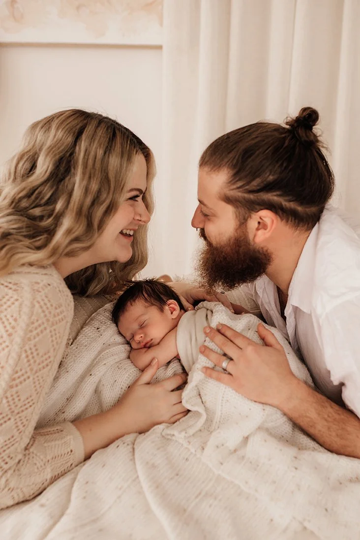New parents smiling at each other while their newborn baby sleeps between them