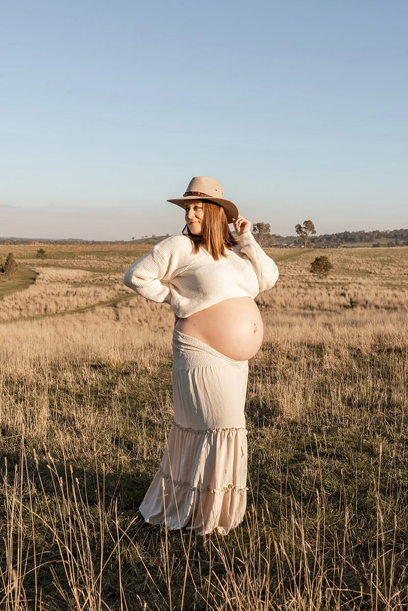 Pregnant woman wearing cowboy hat standing in long grass at sunset in Melbourne's North East