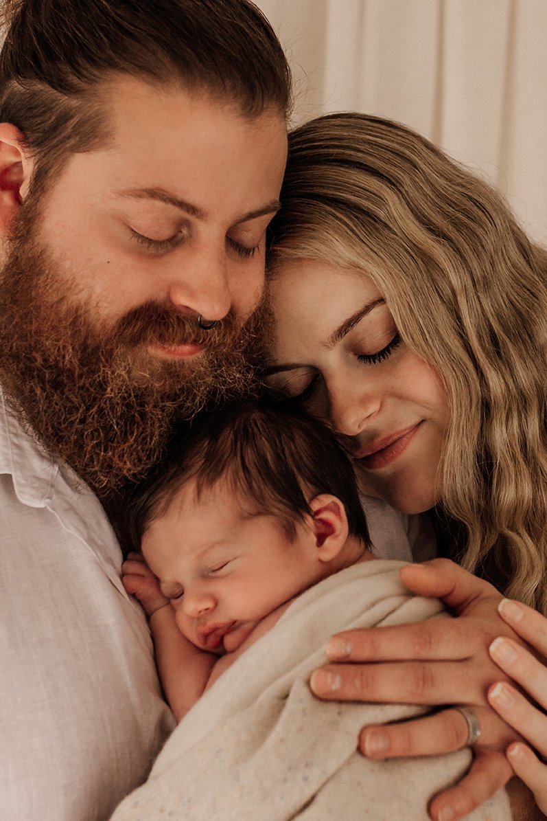Close up image of new parents resting their heads together while newborn baby rests against dad's chest