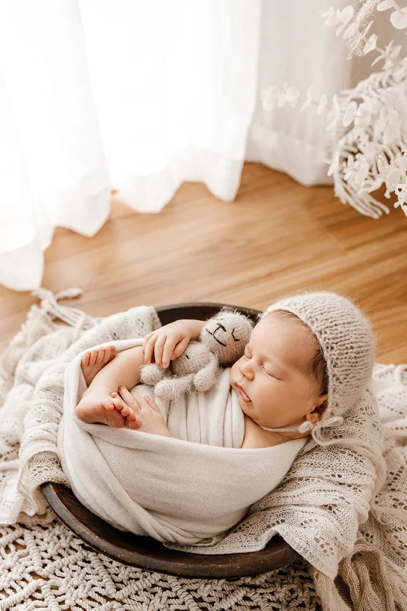 Newborn baby swaddled in a soft blanket, wearing a knitted beanie, cuddling a tiny teddy