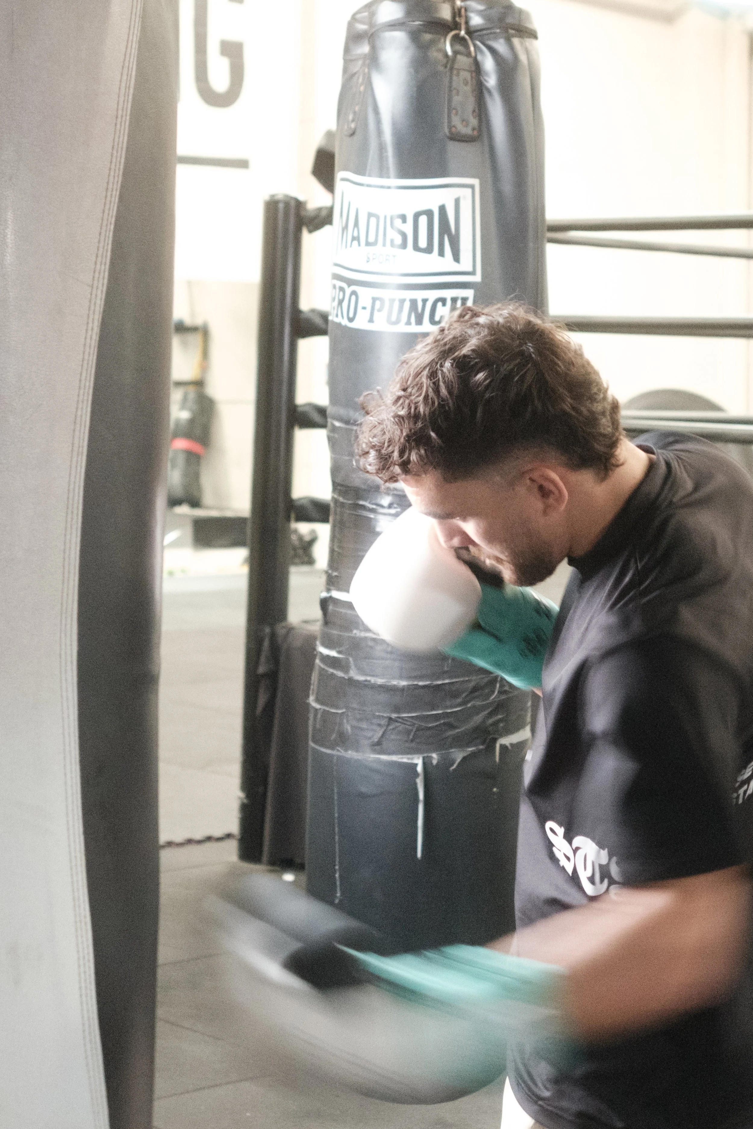 A person with curly hair and a black shirt crouching down in a boxing gym, positioned near a black heavy bag labeled "MADISON SPORT PRO-PUNCH". The person appears to be tying their shoelaces or adjusting something on their foot.