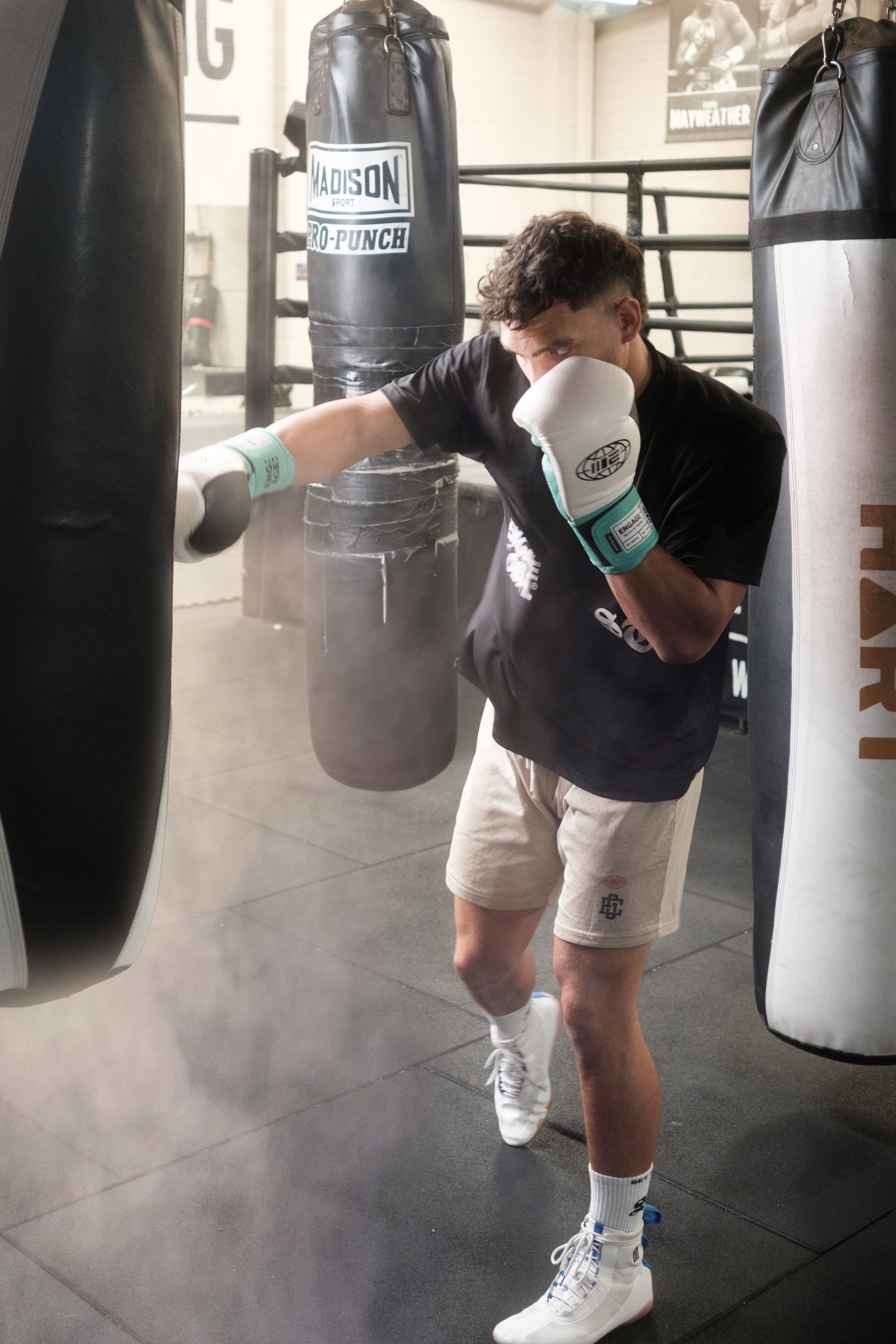Young male boxer practicing punches in a boxing gym, wearing gloves, shorts, and sneakers, with punching bags behind him.