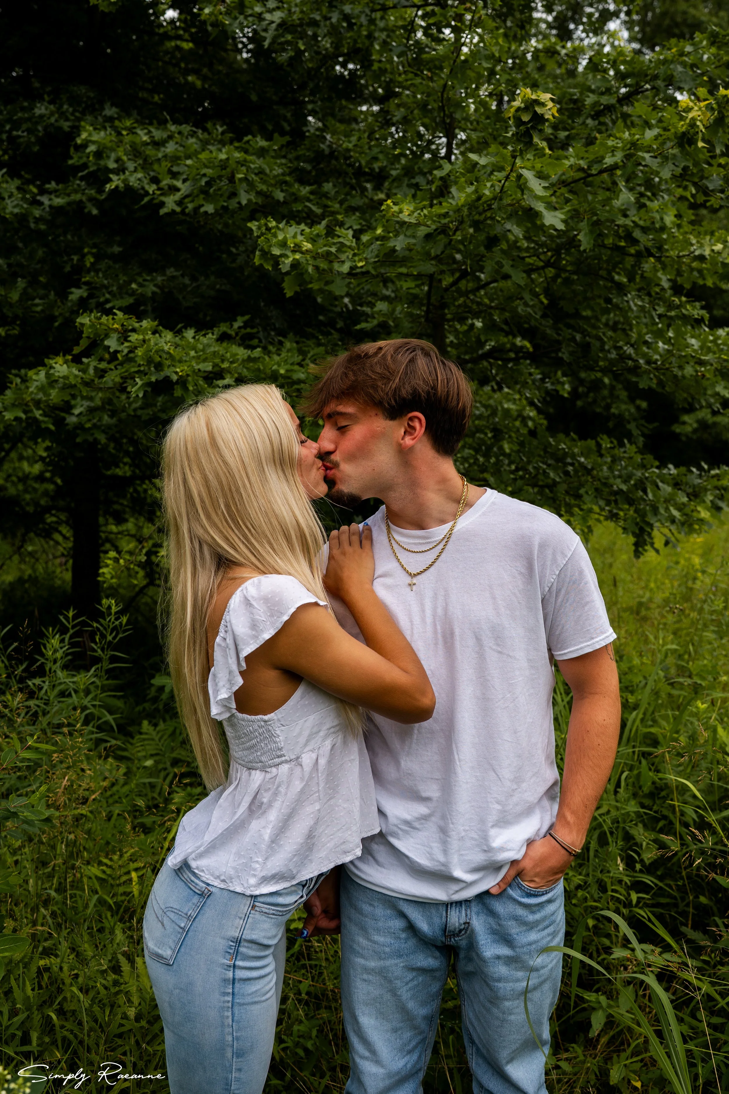 A young couple sharing a kiss outdoors surrounded by green trees and grass.