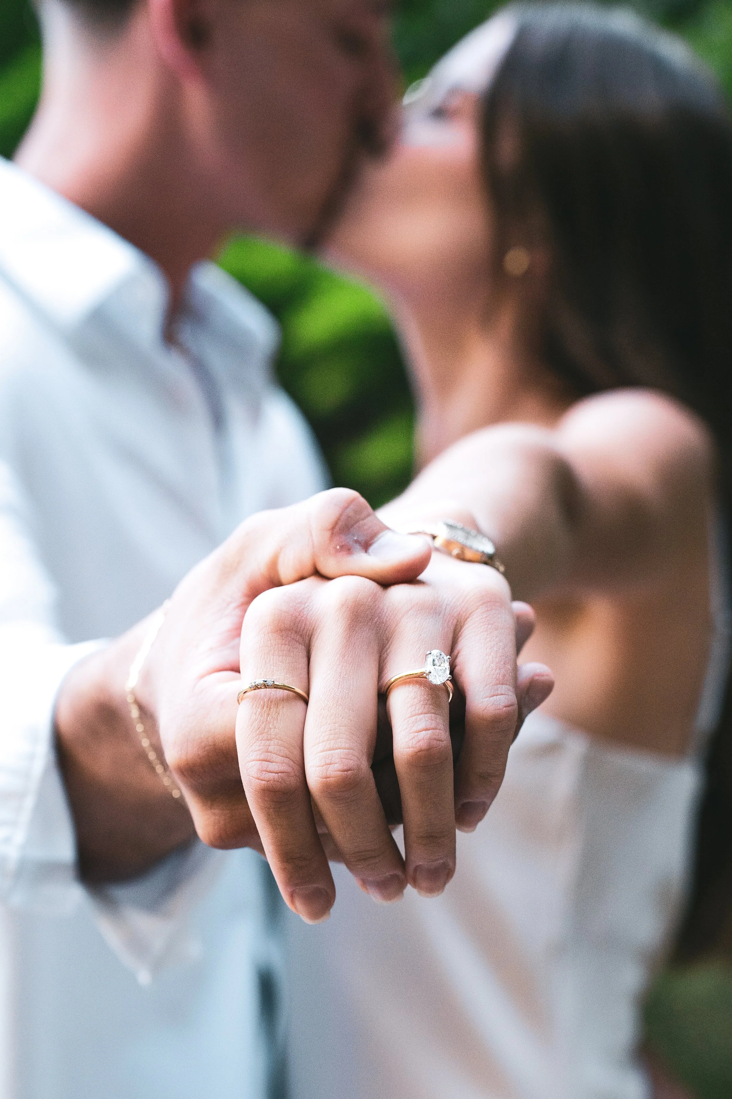 Close-up of a couple's hands showing wedding and engagement rings, with a blurred background of the couple kissing.