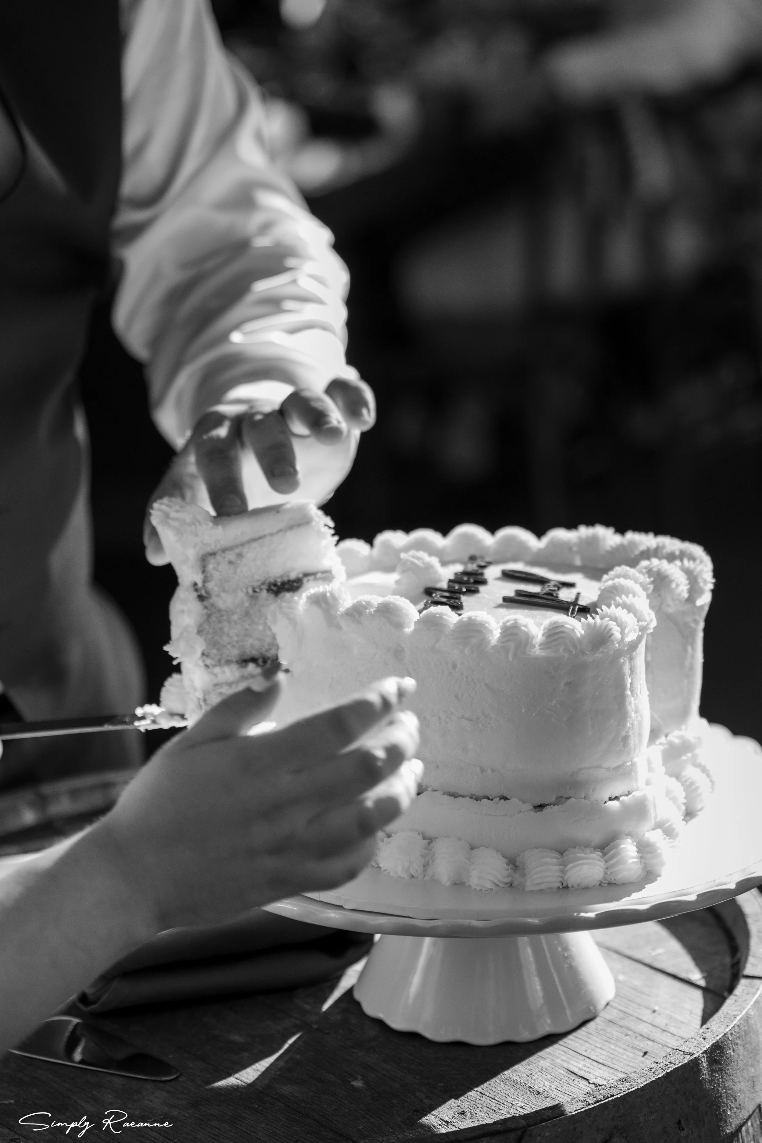 Person cutting a heart-shaped birthday cake with a 'love' decoration on top, on a cake stand, outside.