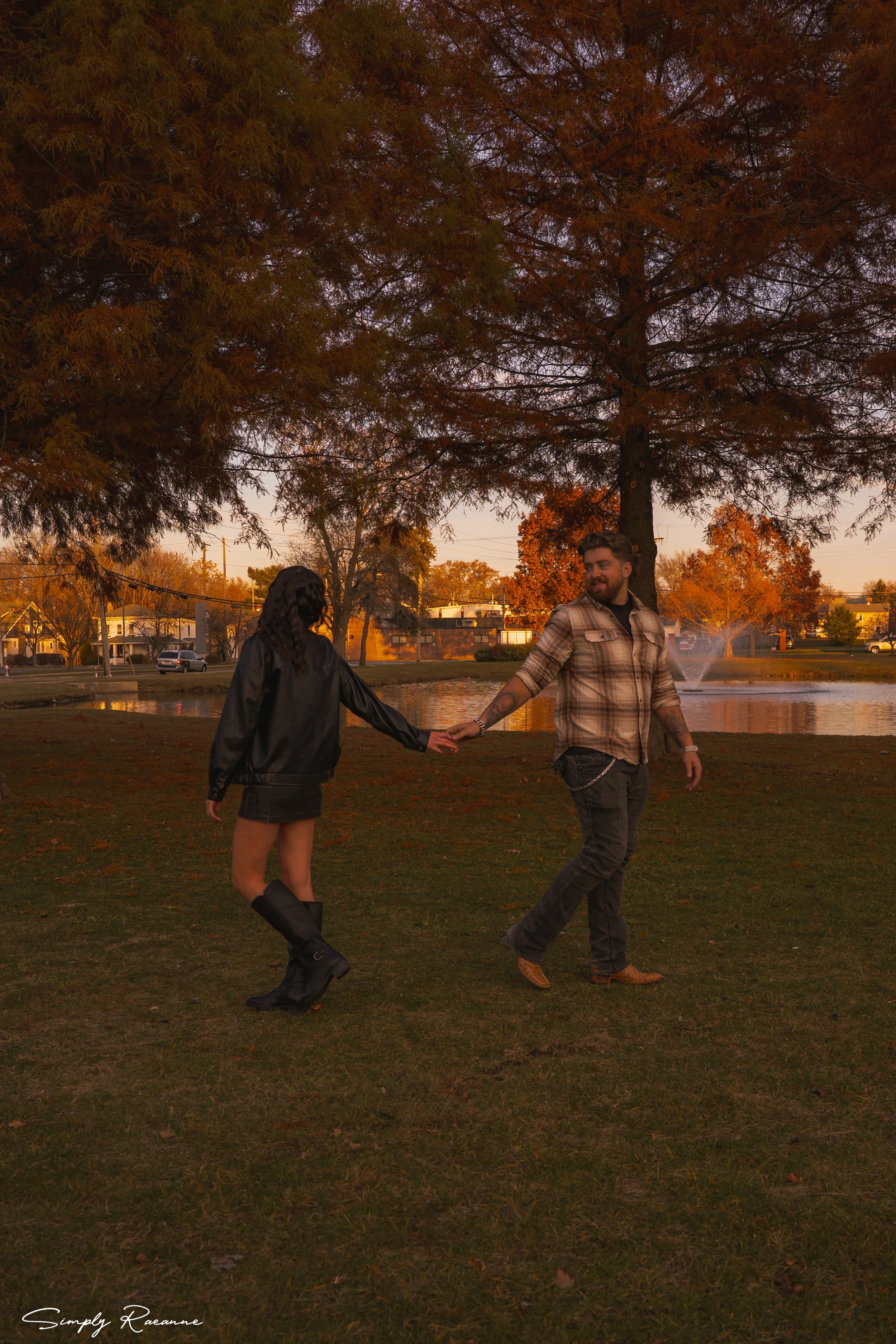 A man and woman holding hands and walking in a park during sunset with fall foliage.