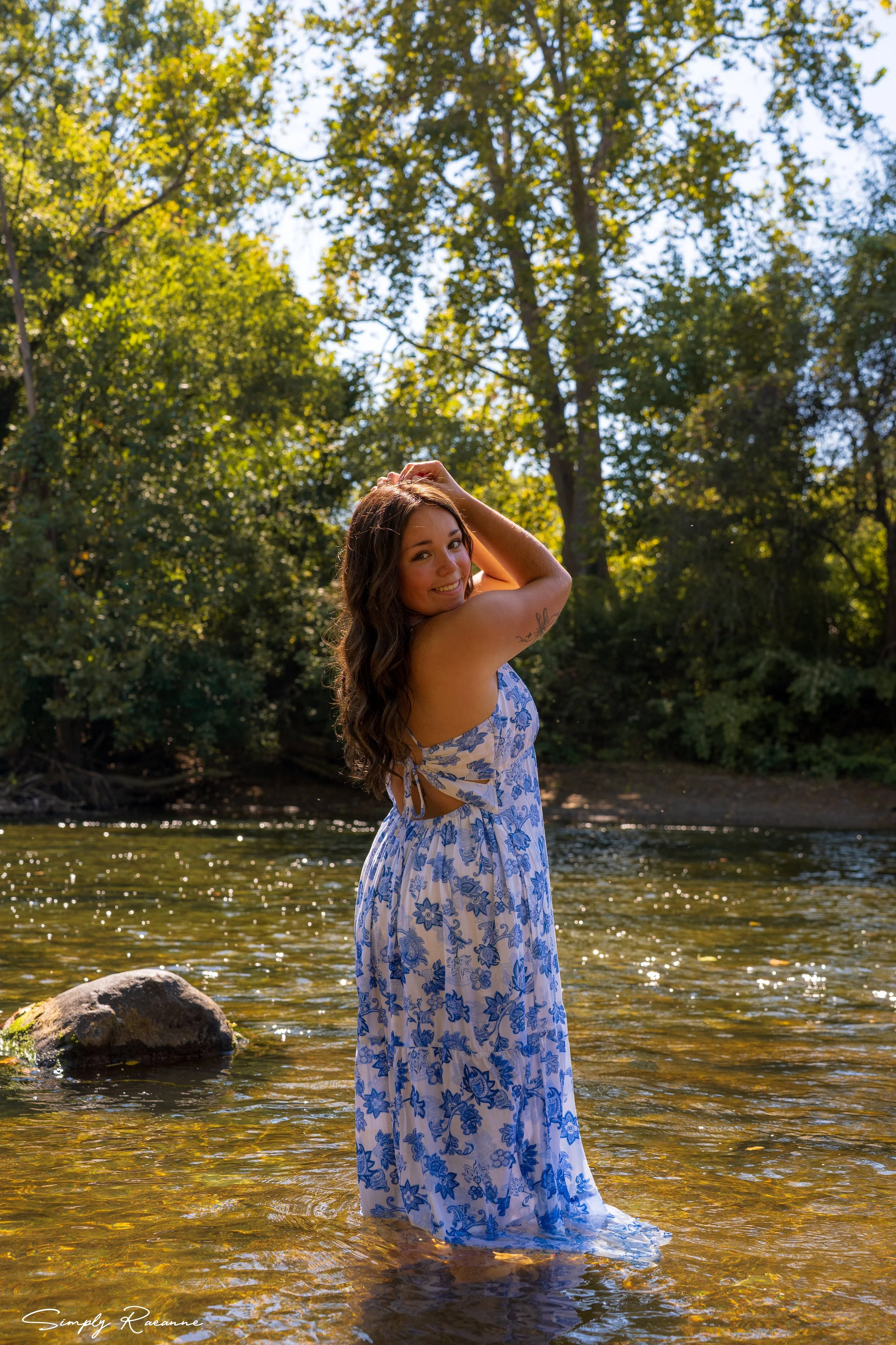 A woman in a blue and white floral dress standing in a shallow river with trees and blue sky in the background, smiling and posing with her hand on her head.