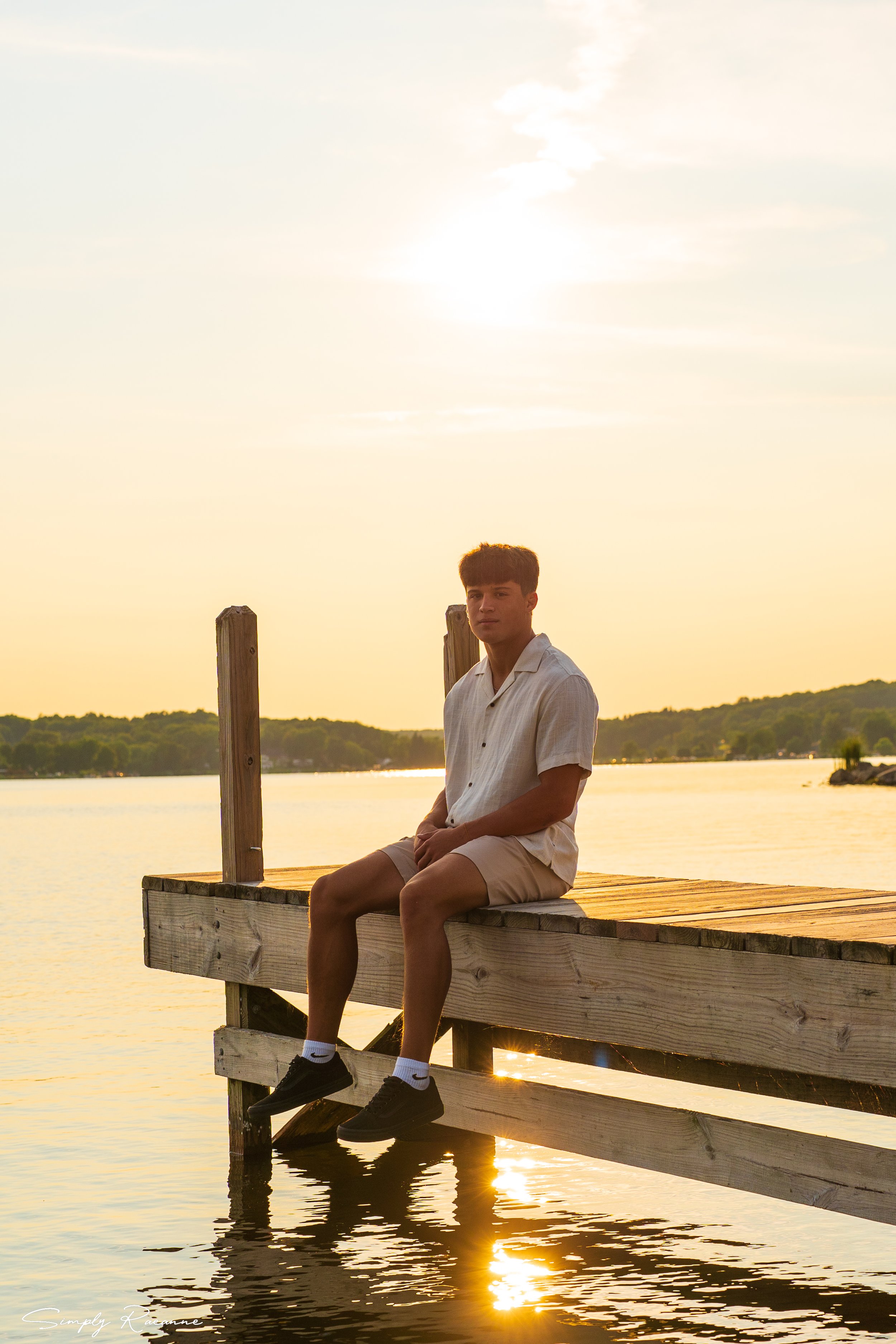 A young man sitting on the edge of a wooden dock by a lake during sunset, with the sun low in the sky and reflecting on the water.
