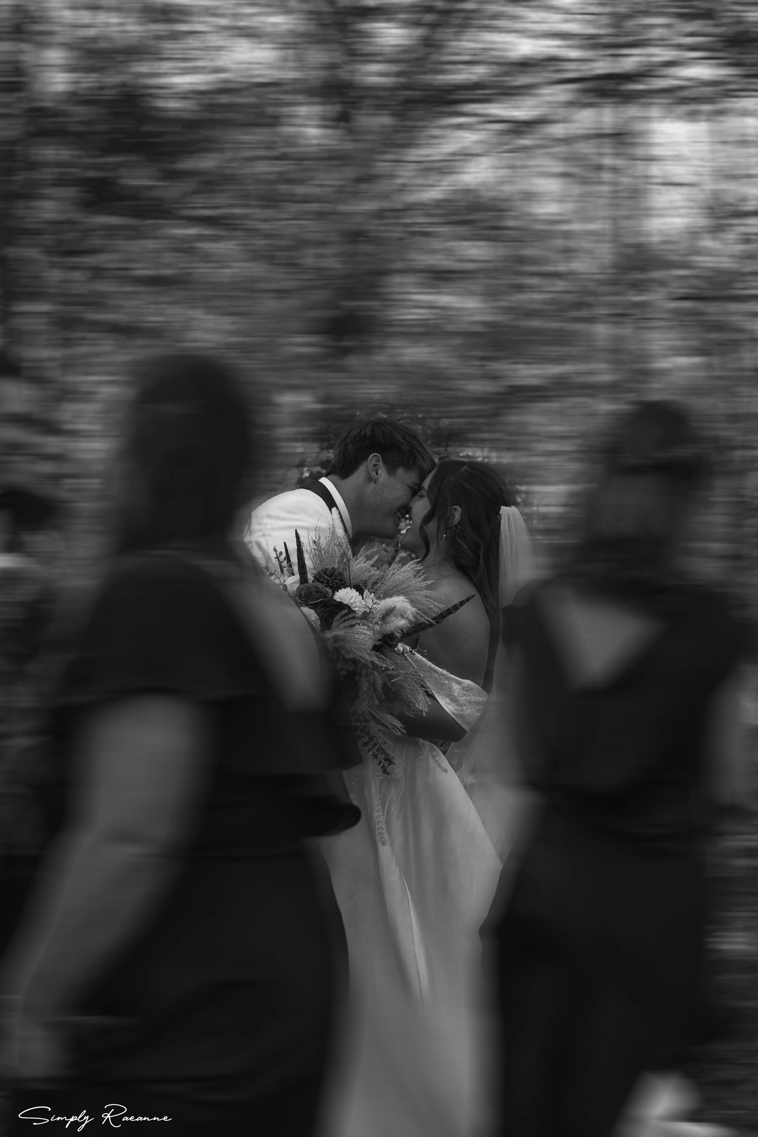 A black-and-white photo of a bride and groom smiling and touching foreheads during their wedding ceremony, with blurred guests in the foreground and a wooded background.