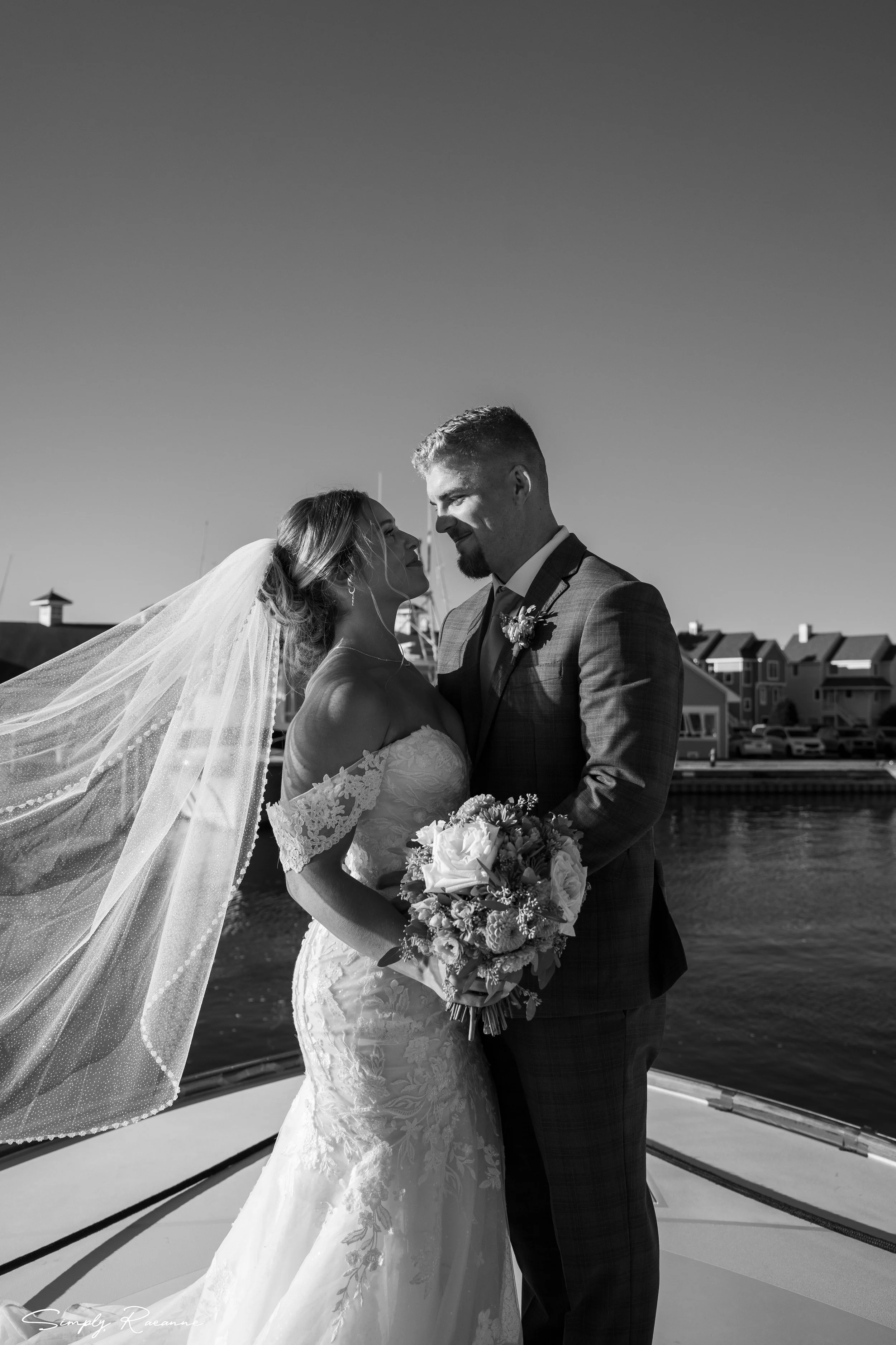 Black and white photo of a bride and groom standing close on a boat dock, facing each other with noses touching, holding a bouquet of flowers, with waterfront and houses in the background.