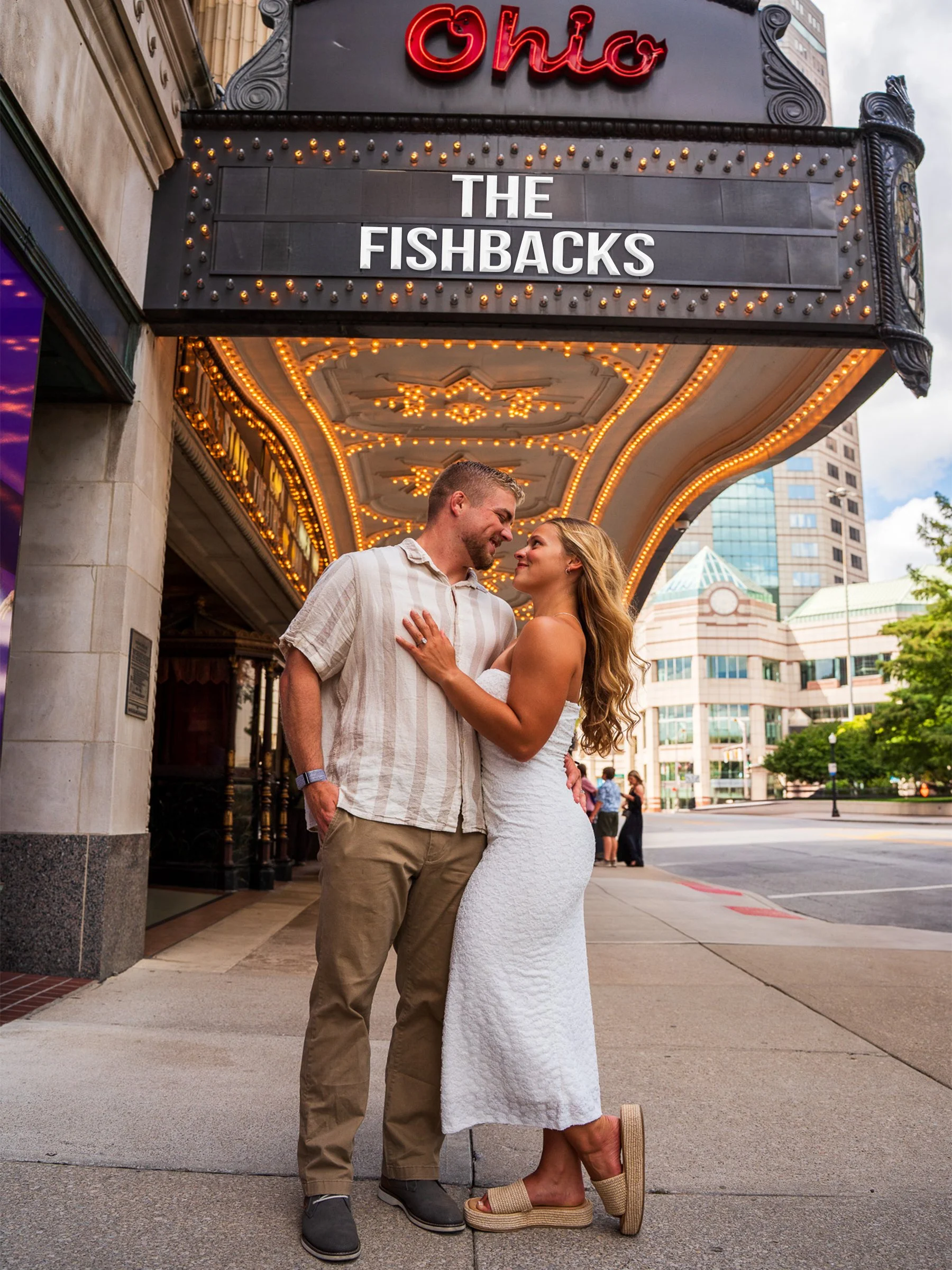 A couple in casual summer clothes standing close and smiling in front of a vintage theater marquee with the sign "The Fishbacks" and red neon letters saying "Olio". The woman is wearing a strapless white dress and beige sandals, while the man is in a