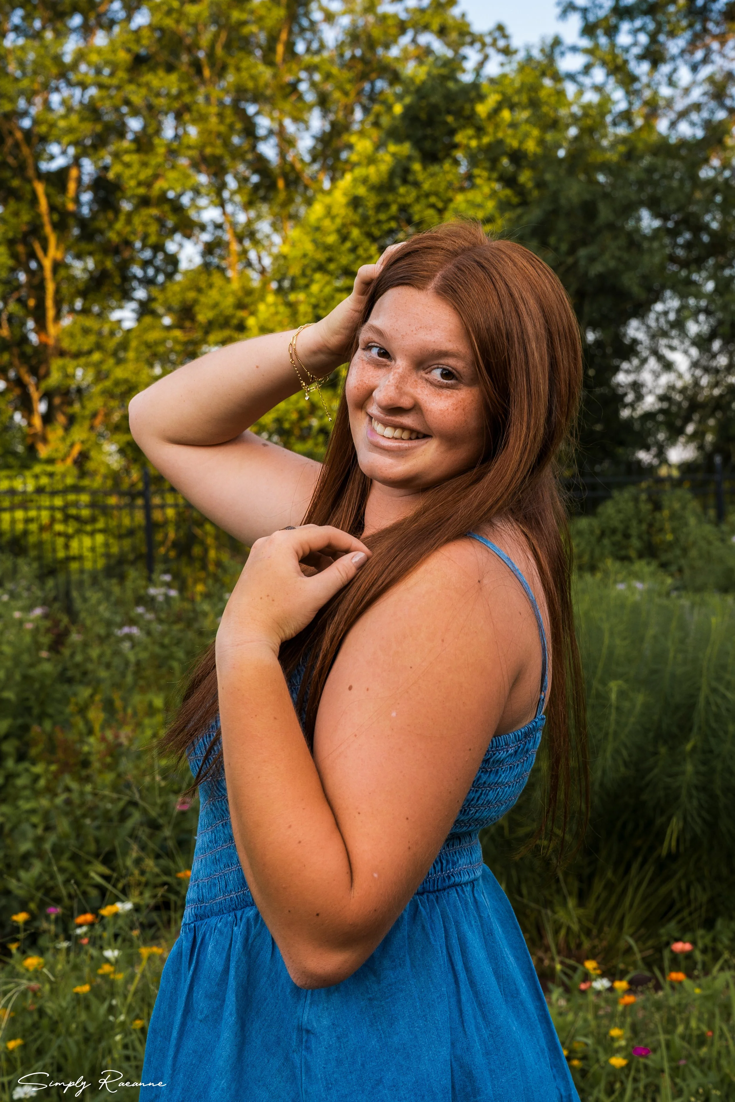 A young woman with long red hair, smiling and looking at the camera, stands in a garden with blooming flowers and trees in the background during golden hour.