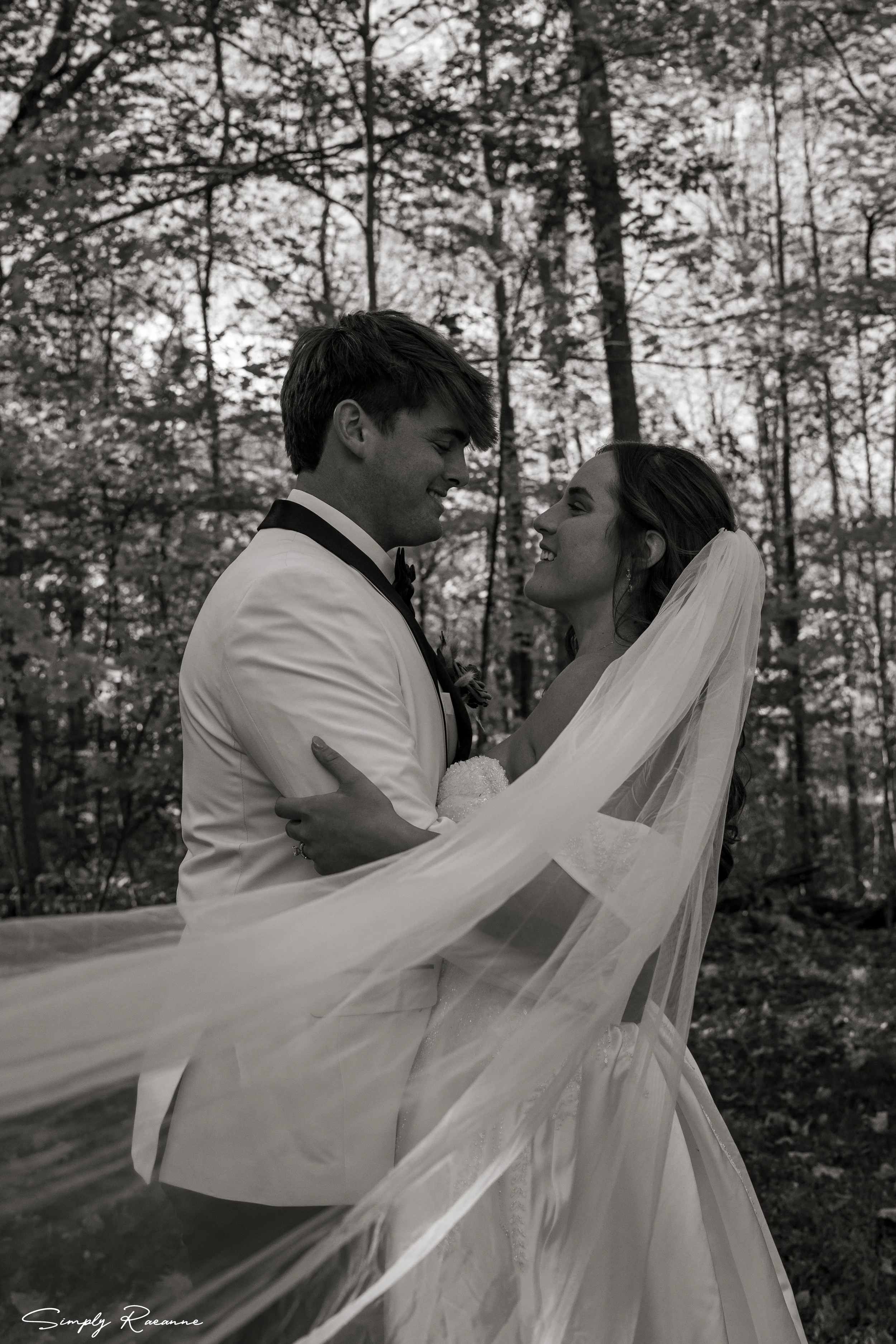 A bride and groom embrace outdoors in a wooded area, smiling at each other, with the bride wearing a veil and the groom in a tuxedo, captured in black and white.