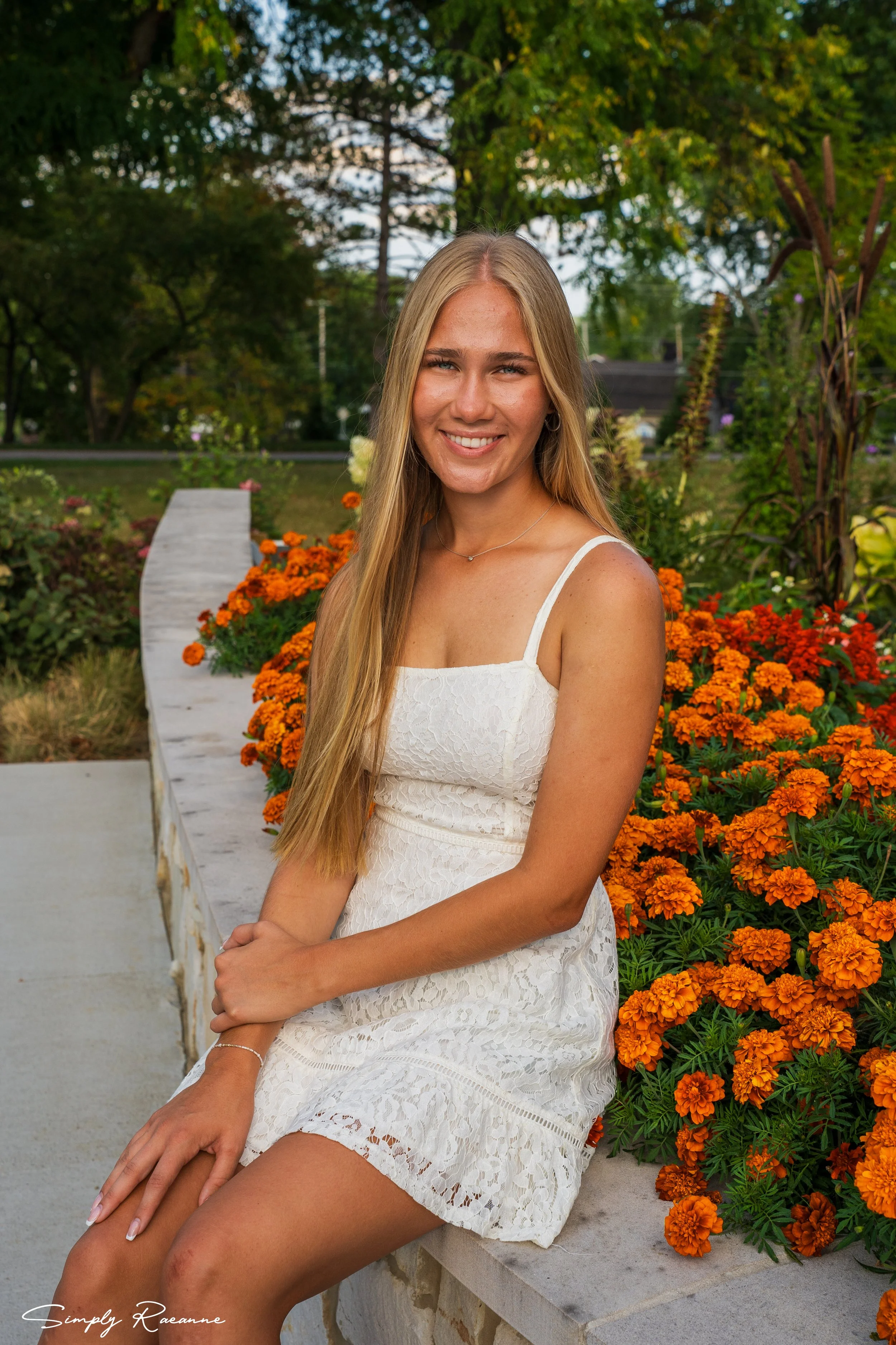 A young woman with long blonde hair in a white lace dress sitting on a stone ledge beside orange marigold flowers in a garden.