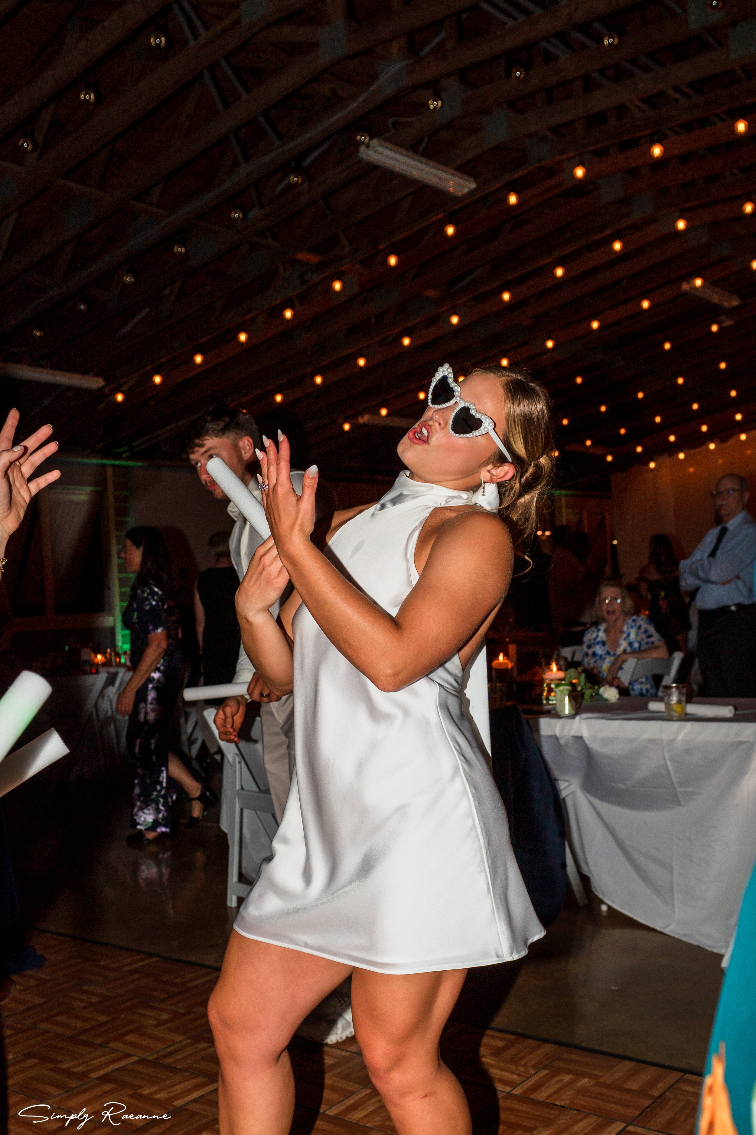 Woman wearing sunglasses and a satin dress dancing at a party, with other guests in the background and a decorated ceiling.