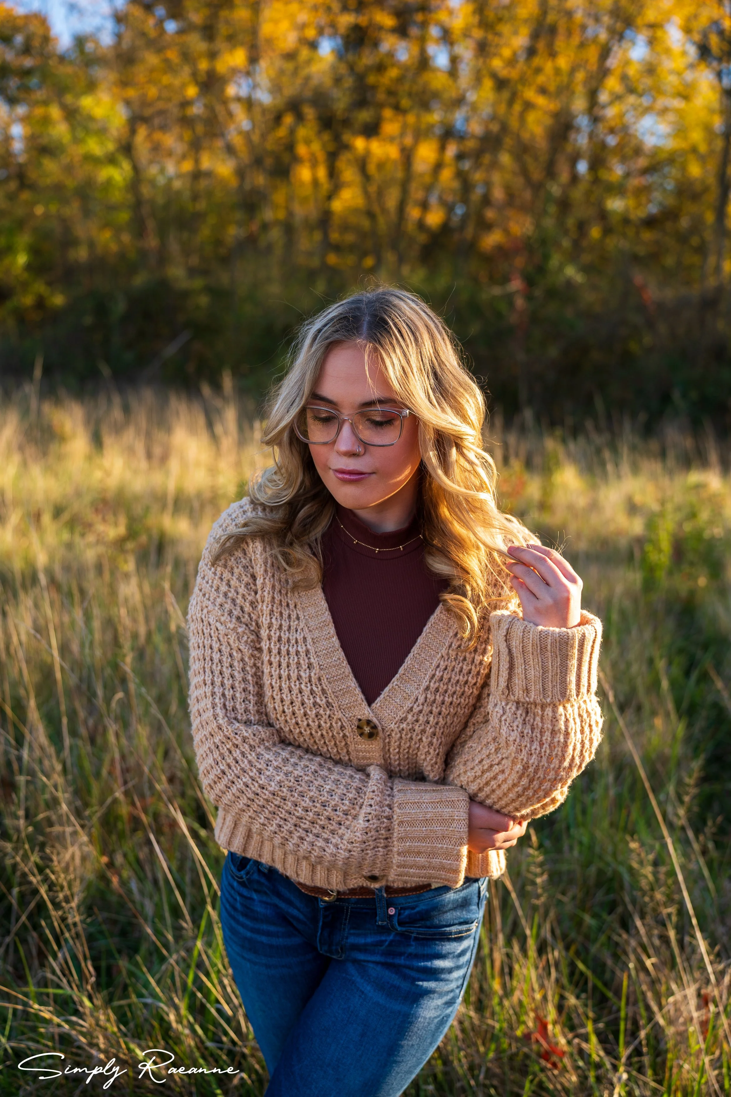 A young woman with blonde curly hair, glasses, and light makeup is standing outdoors in a field with tall grass and autumn trees. She is wearing a beige knitted cardigan, a maroon top, and blue jeans, with her eyes closed and one hand touching her hair. She is illuminated by warm sunlight during sunset or late afternoon.