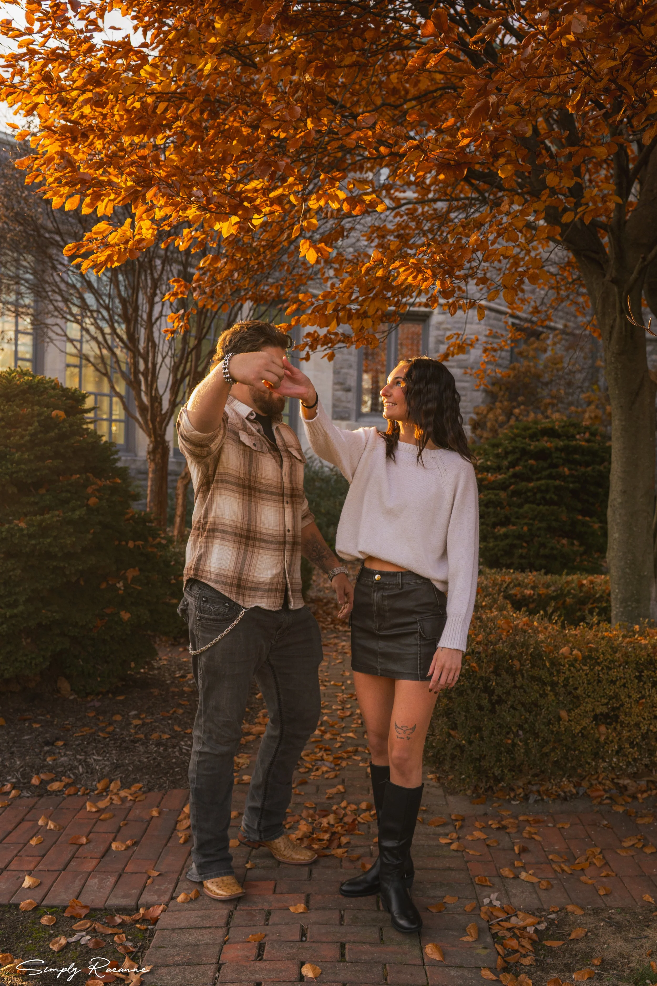 A man and woman dancing outdoors under orange autumn leaves, smiling at each other, during sunset.