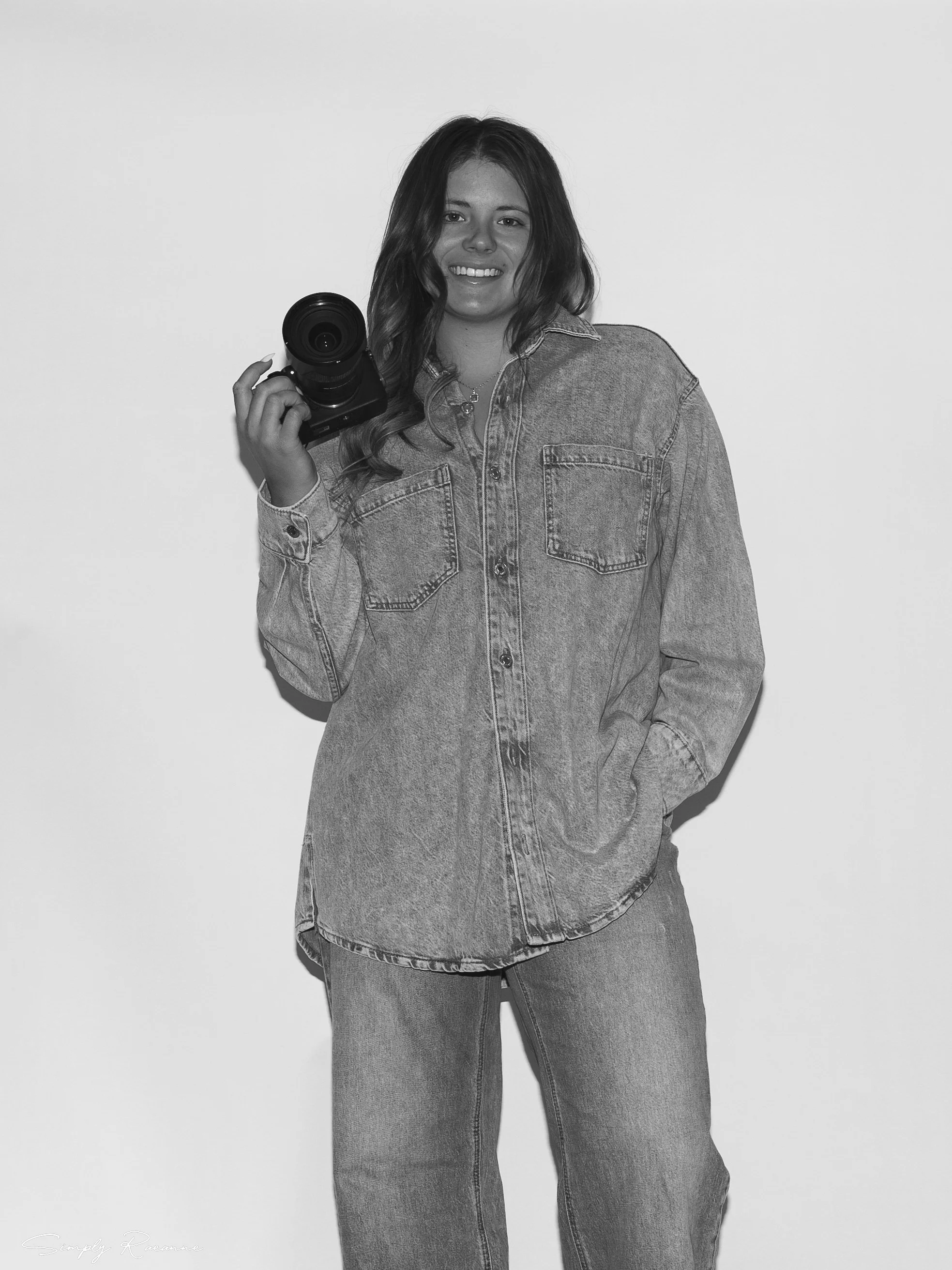 A woman standing against a plain background, holding a camera in her right hand, wearing a denim jacket and jeans, smiling at the camera.