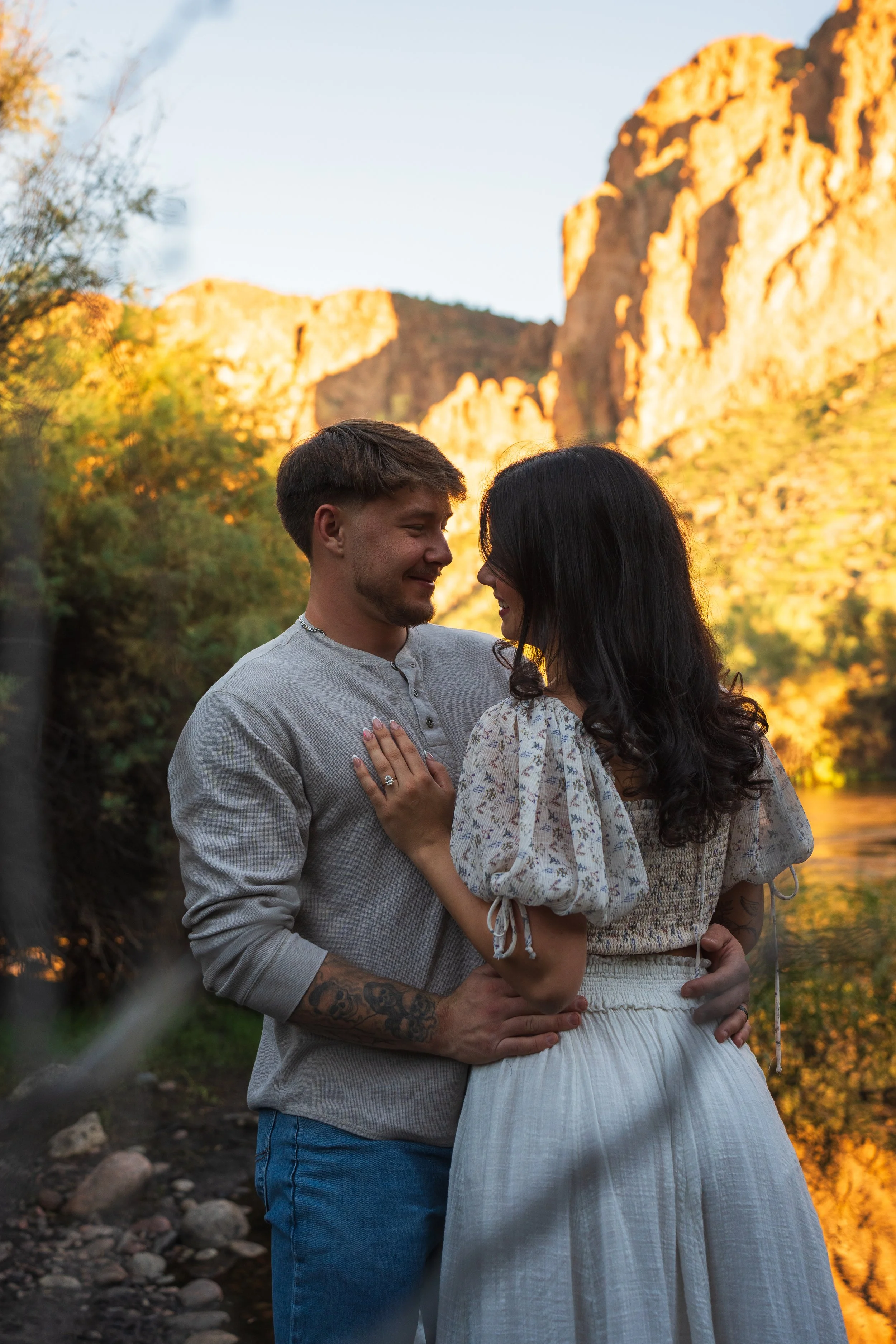 A couple standing close together outdoors at sunset, with mountains in the background and a river nearby.