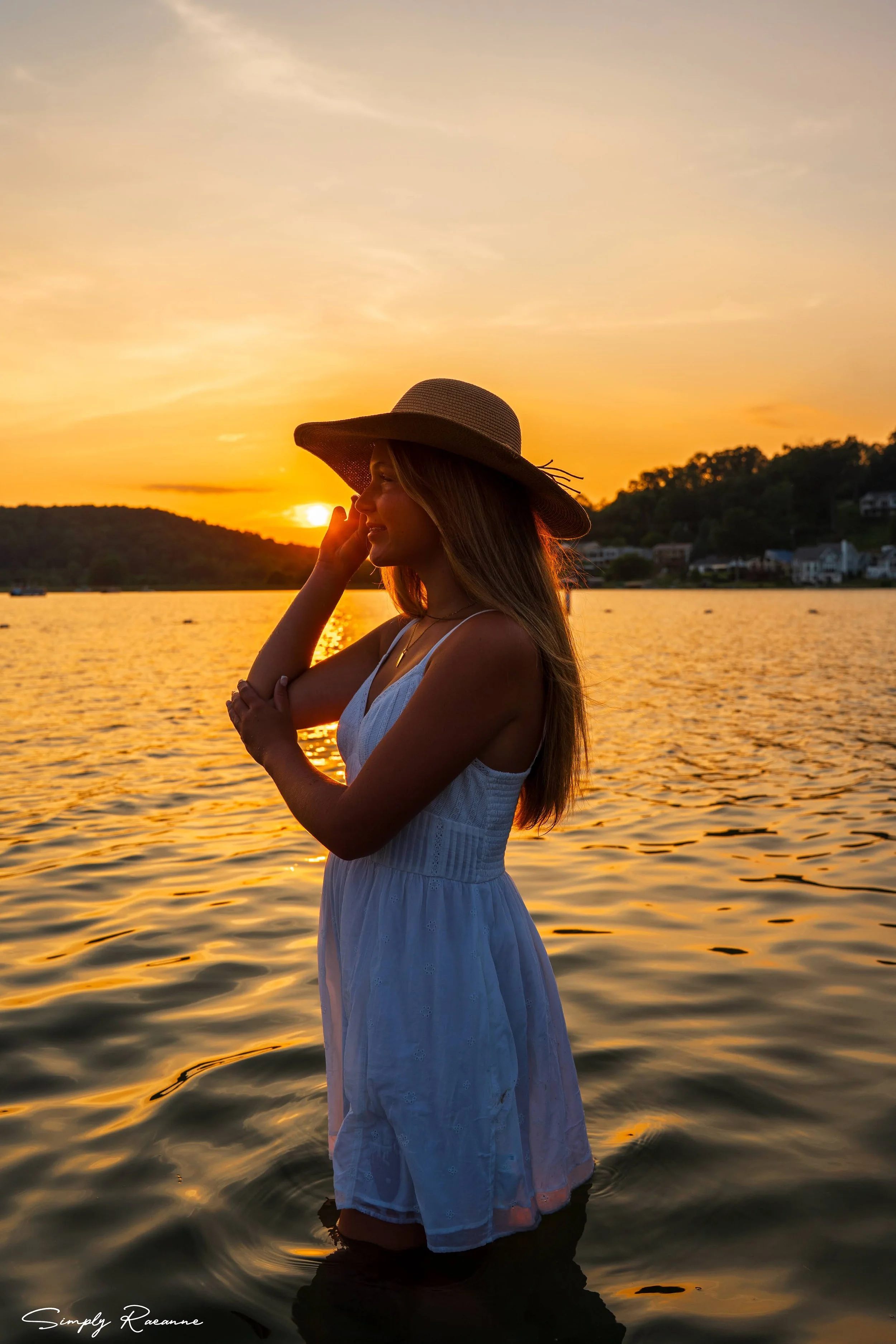 A woman wearing a wide-brimmed hat and a white dress standing in a body of water at sunset, with hills and houses in the background.