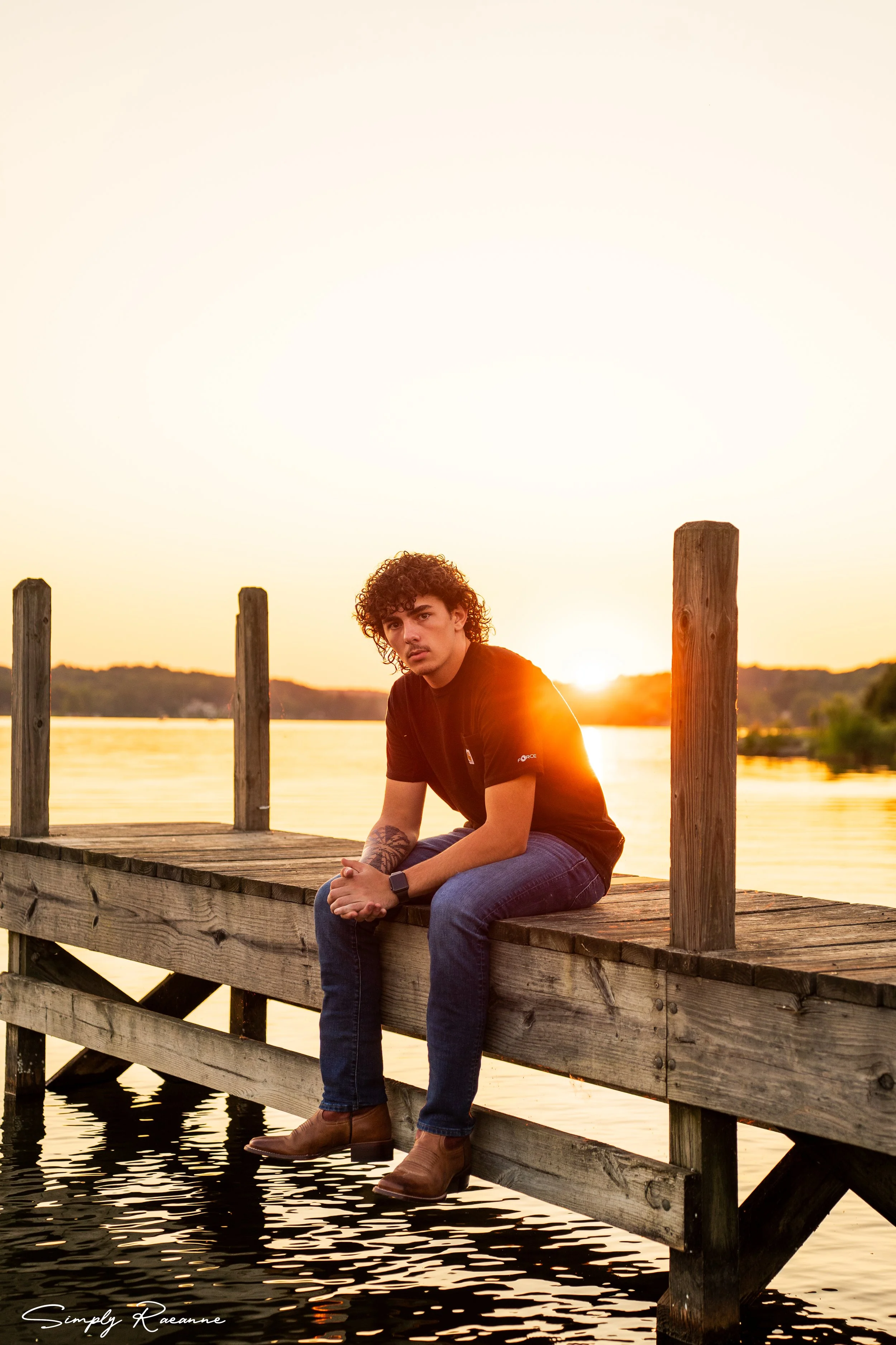 A young man with curly hair sitting on a wooden dock at sunset by a lake, looking at the camera, with water underneath and a landscape in the background.