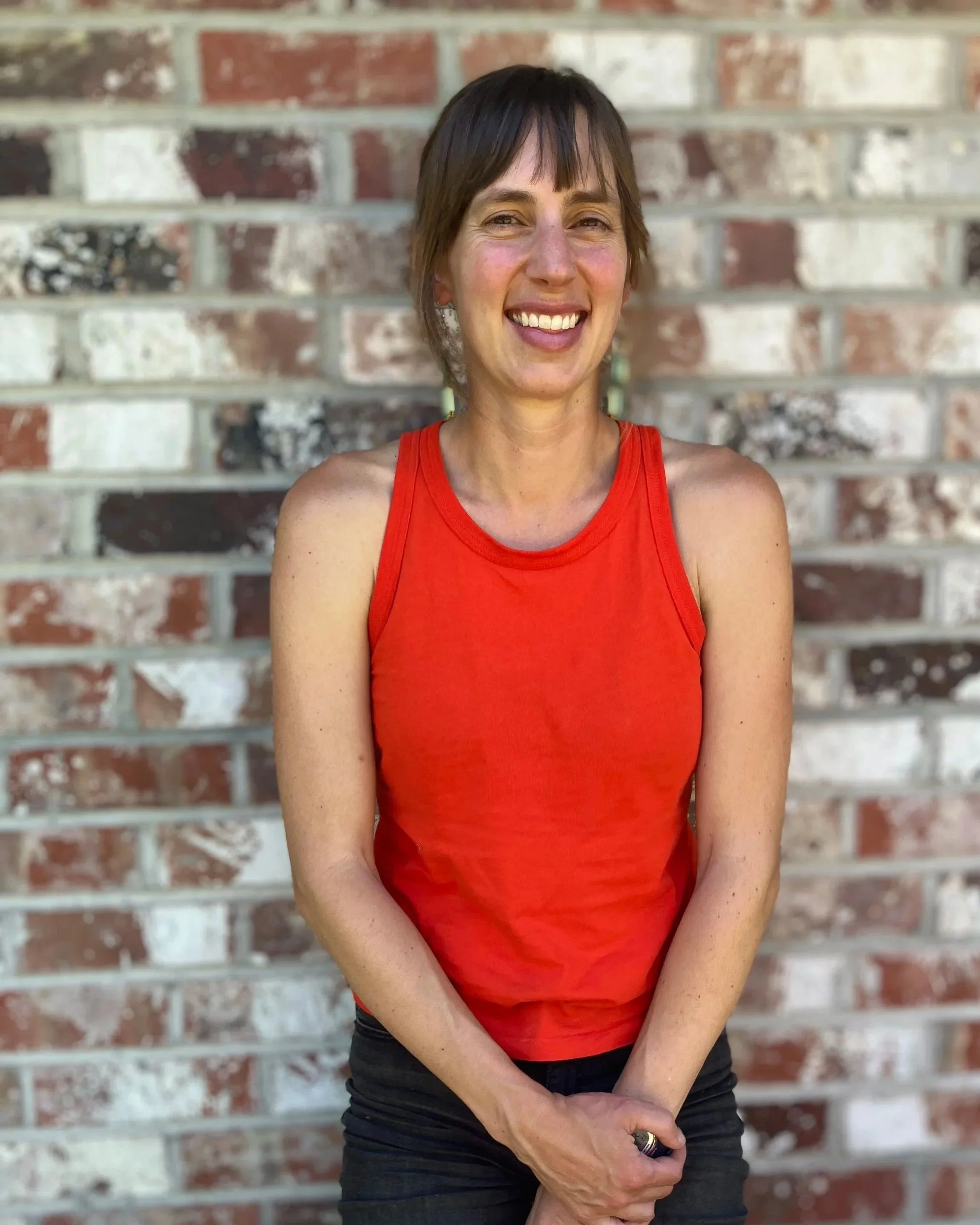 A woman with short brown hair wearing a red tank top and black pants, standing in front of a colorful brick wall, smiling at the camera.