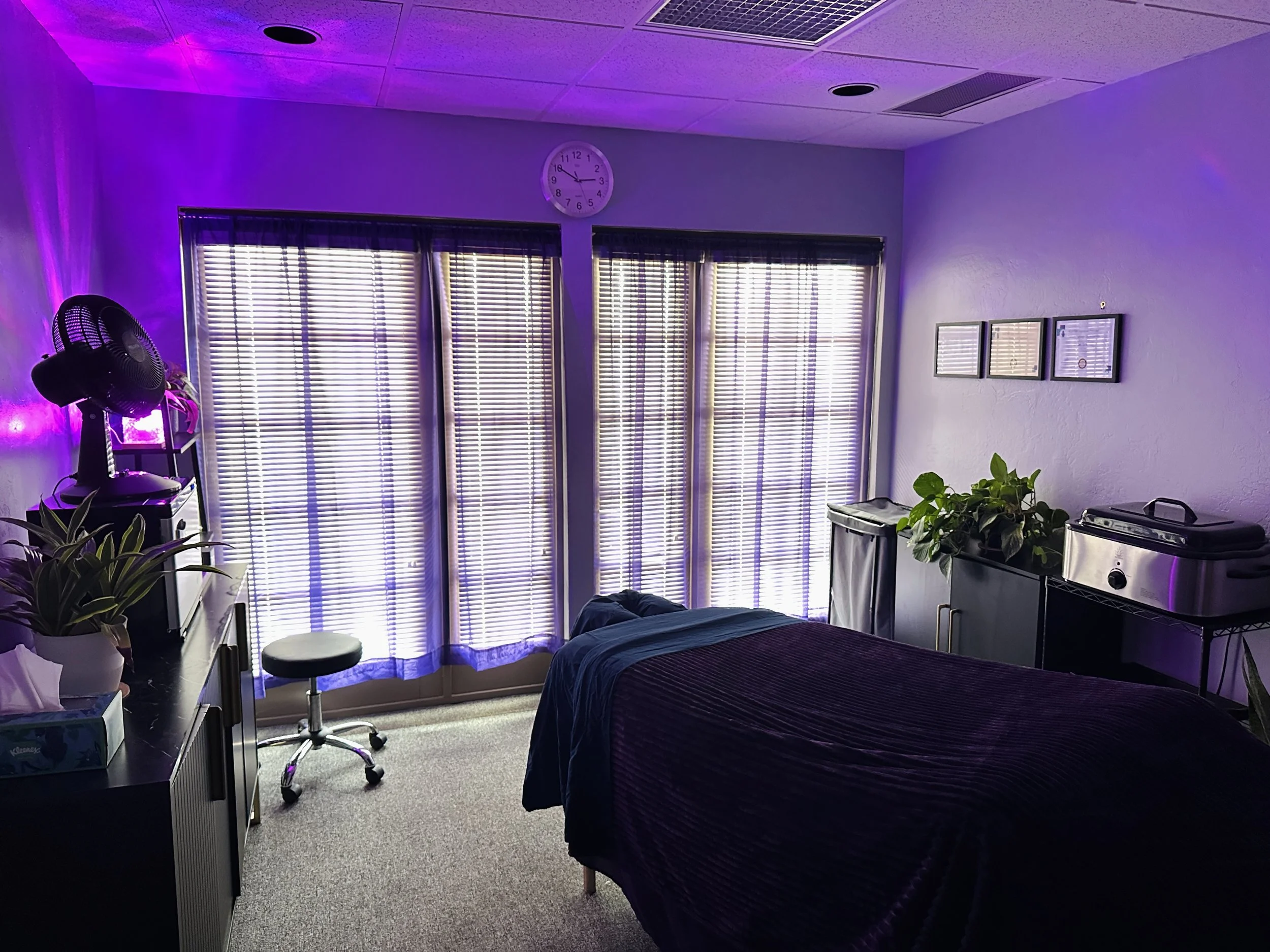 Massage studio treatment room with purple lighting, window blinds, plants, and a comfortable massage table.