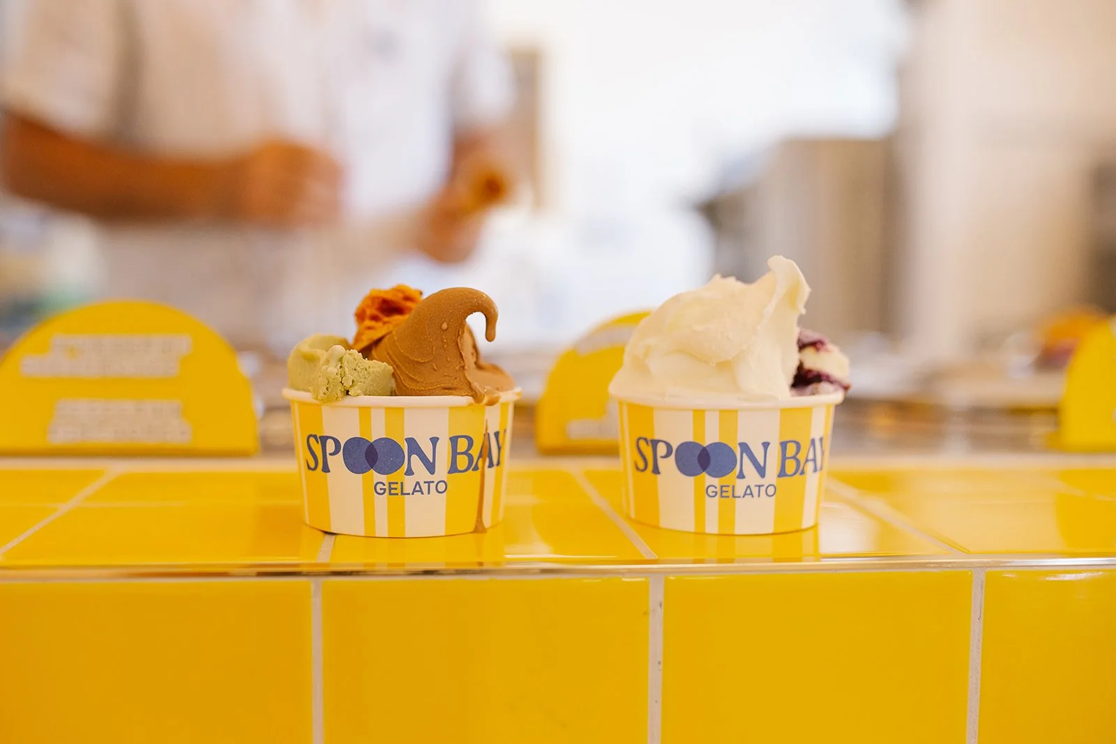 Two cups of gelato on a yellow tiled surface with a yellow and white striped pattern, featuring the words 'Spoon Bay Gelato' and a logo with a heart shape, with blurred background of a person and a shop setting.