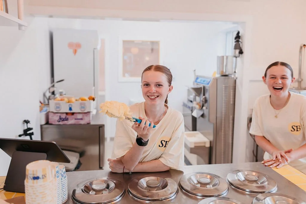 Two young women in a bake shop, one holding a spatula with frosting, the other laughing, surrounded by baking equipment.