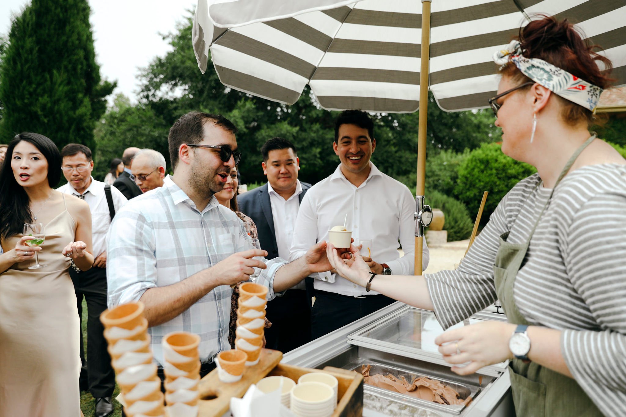 A group of people at an outdoor gathering, enjoying ice cream served from a vendor under a striped umbrella.