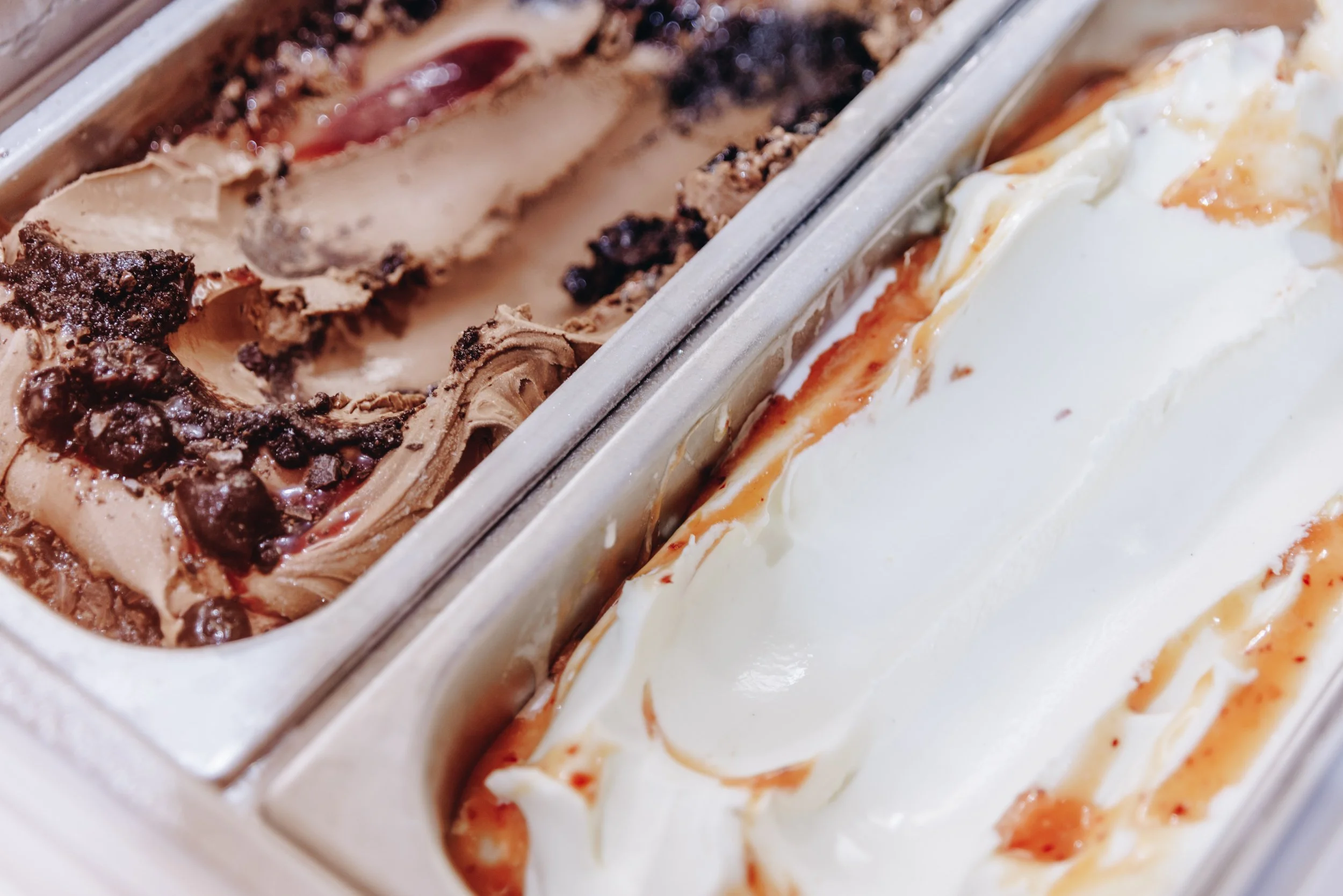 Close-up of two flavors of ice cream in a metal freezer: chocolate with chocolate chunks and caramel swirls, and vanilla with caramel drizzle.