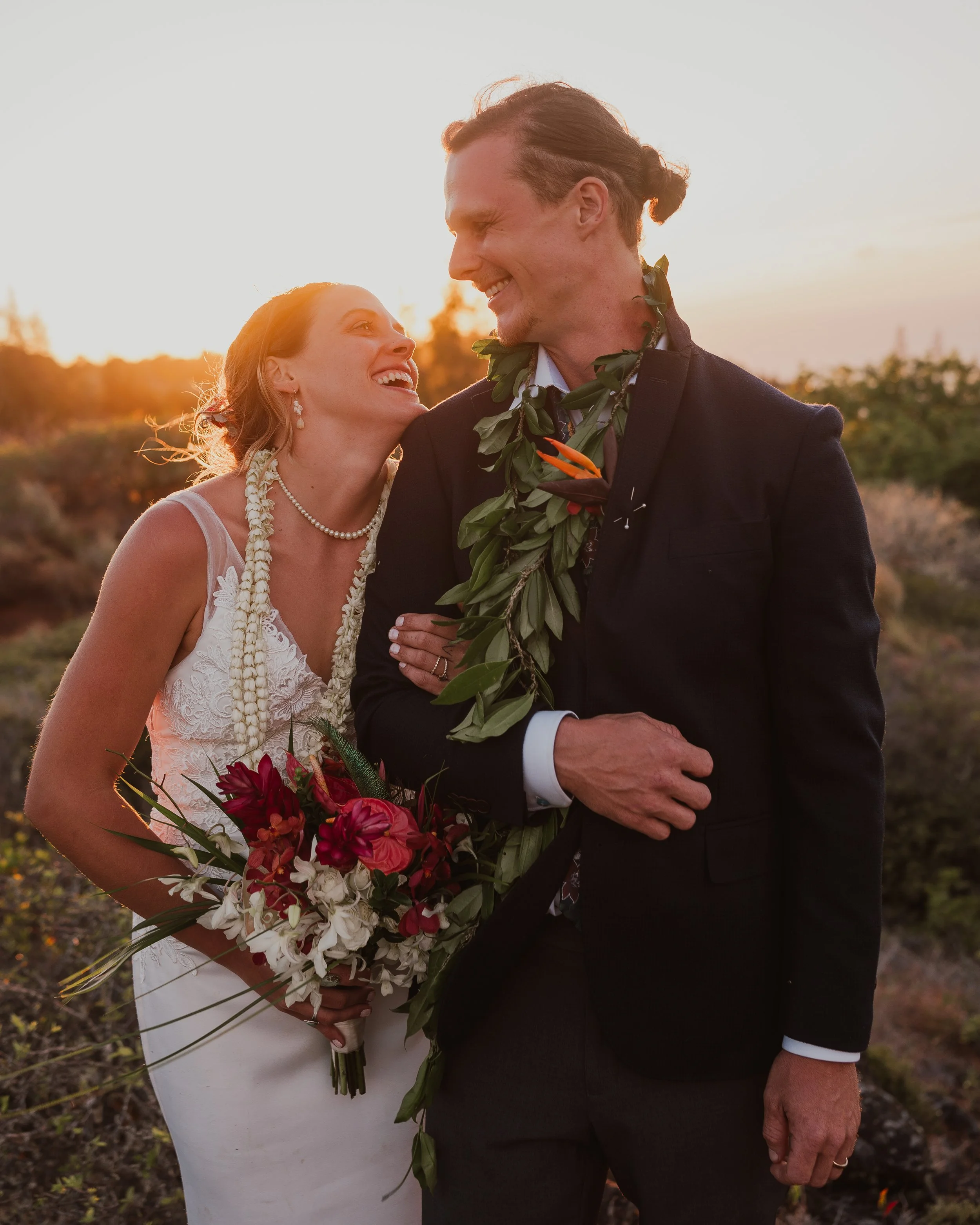 A happy bride and groom smiling at each other during their outdoor wedding at sunset, with the bride holding a bouquet of red and white flowers and the groom wearing a lei of greenery.
