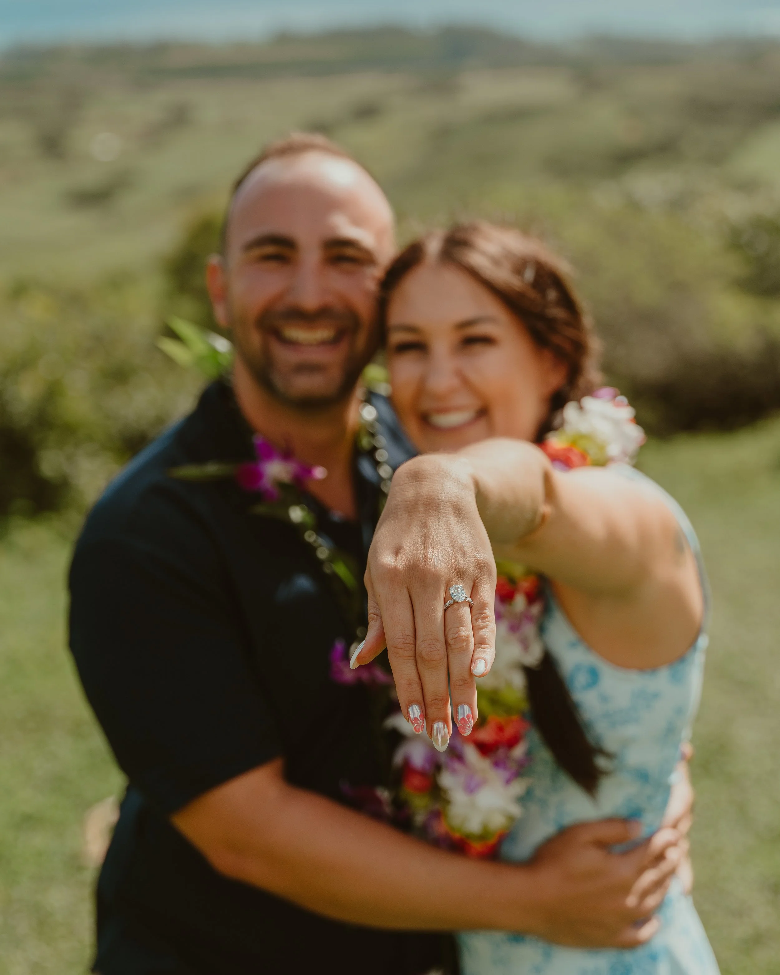 Couple showing off engagement ring during outdoor celebration, smiling, with a blurred natural background.