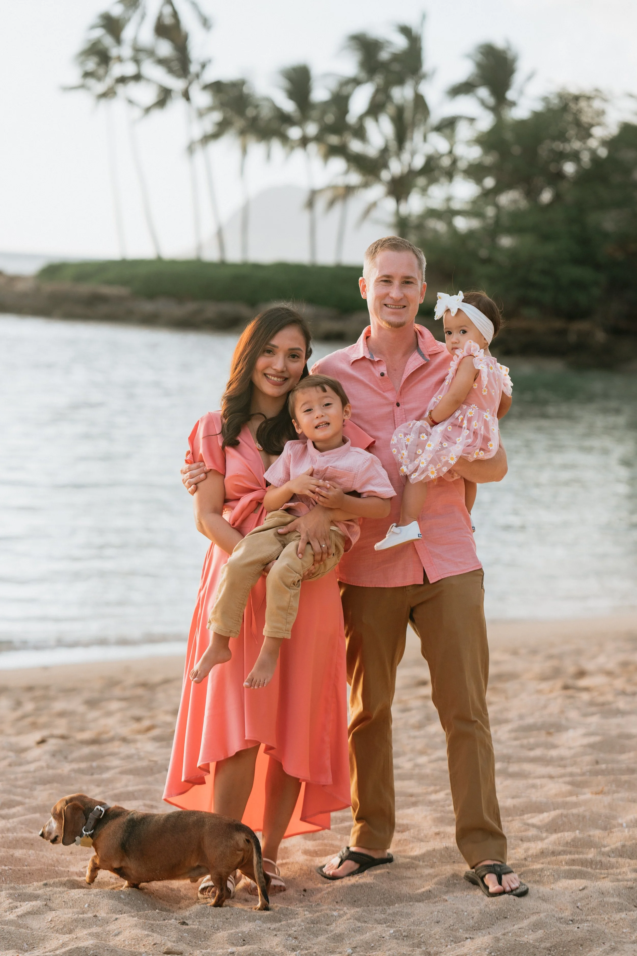 A family of four with a dog at the beach, smiling and dressed in pink and neutral colors, with palm trees and water in the background.