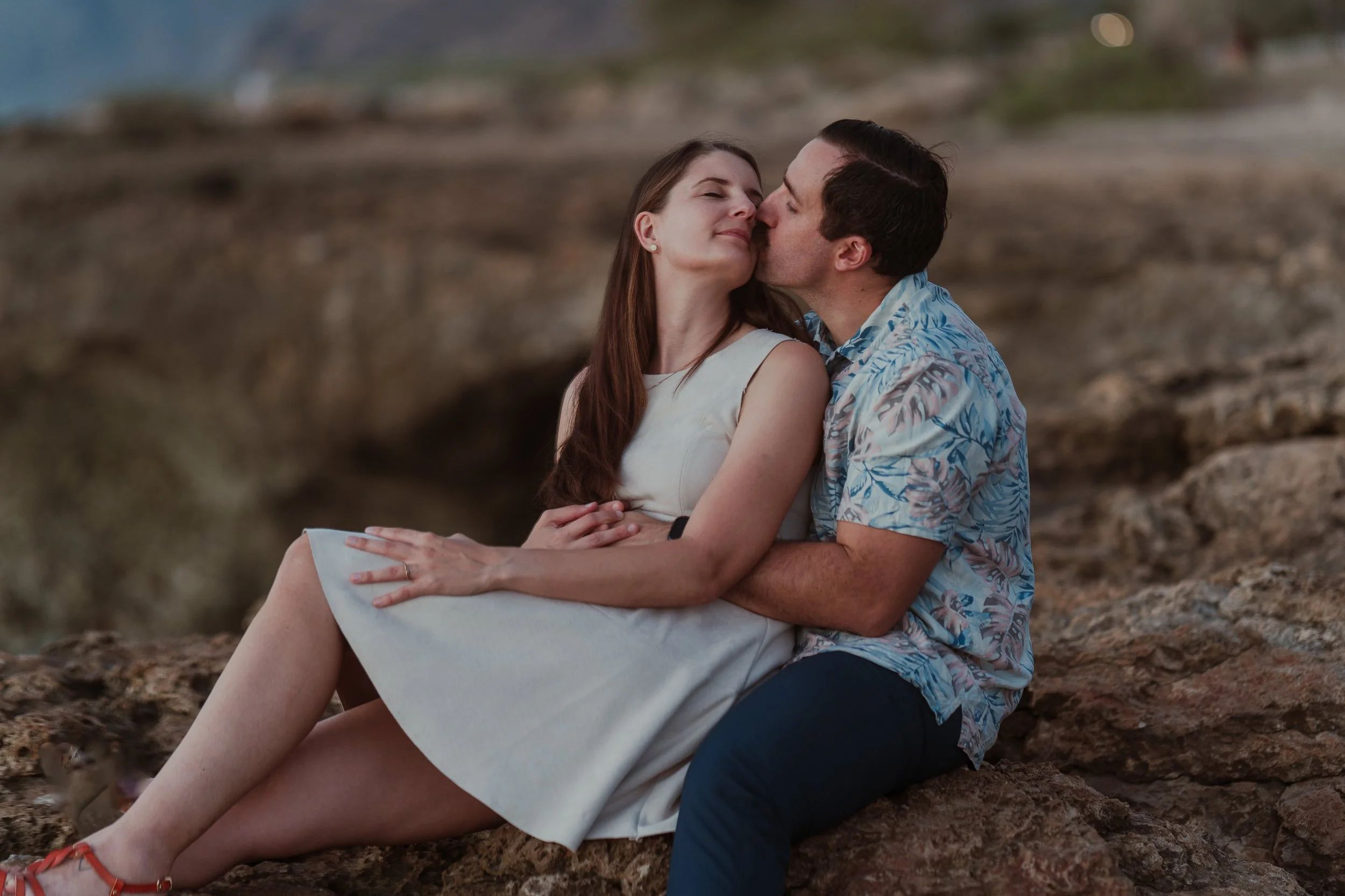 A couple sitting on rocks outdoors, kissing and smiling, with a blurred natural background at dusk.