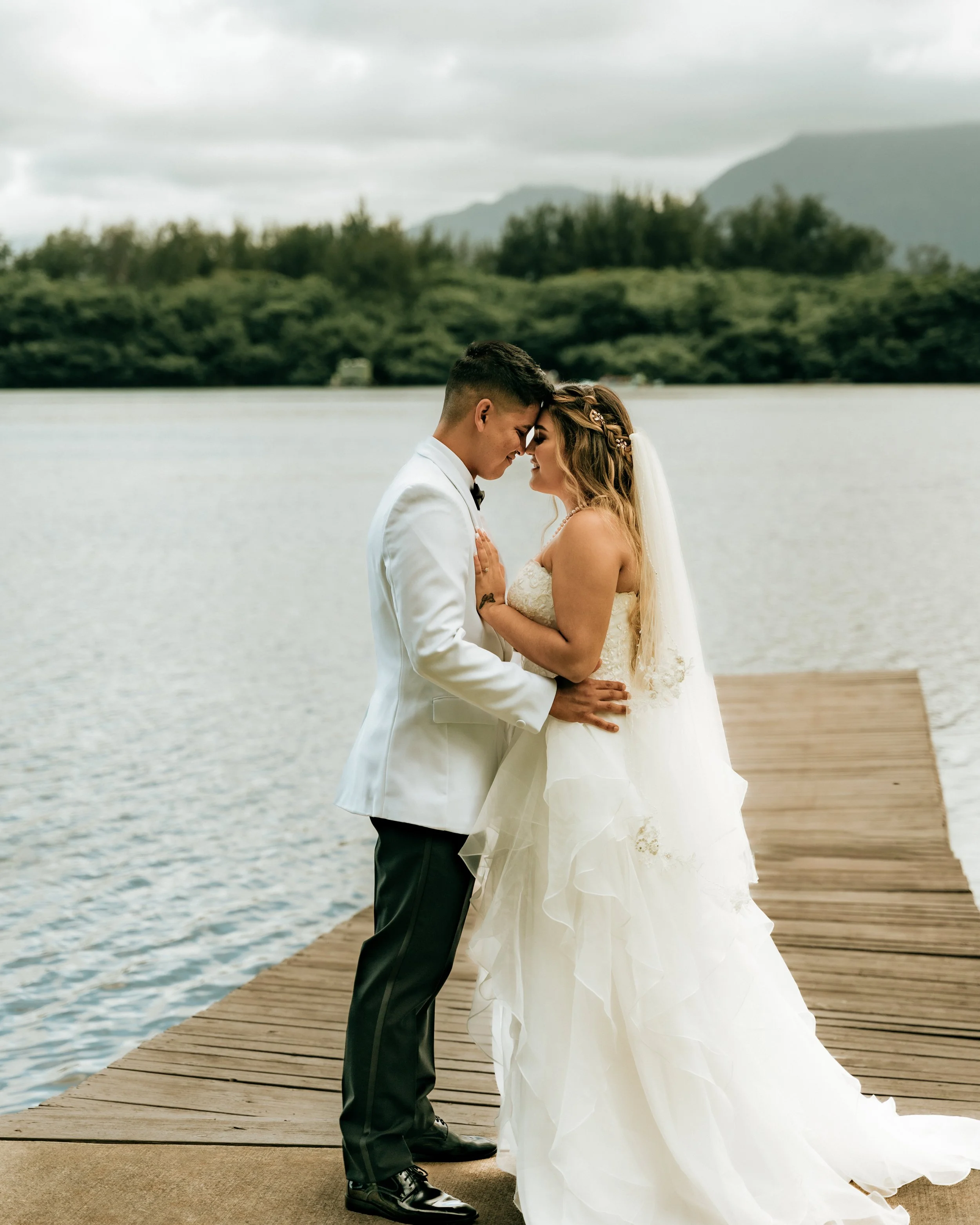 A newlywed couple, dressed in wedding attire, standing close together on a wooden dock by a lake outdoors, with a backdrop of trees and mountains under a cloudy sky.
