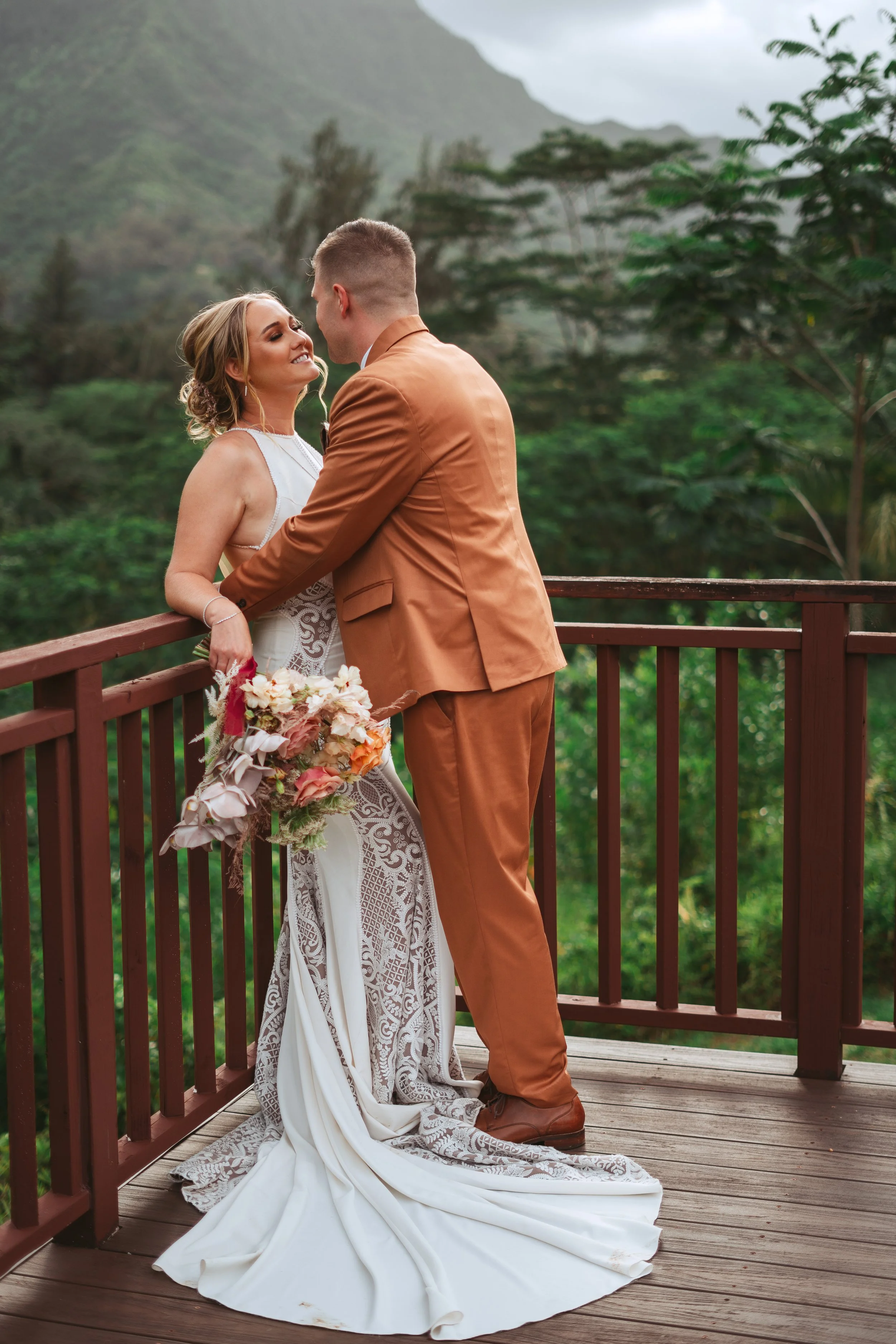 A bride and groom on a wooden balcony with green mountains in the background, embracing and smiling at each other. The bride is holding a bouquet of flowers.