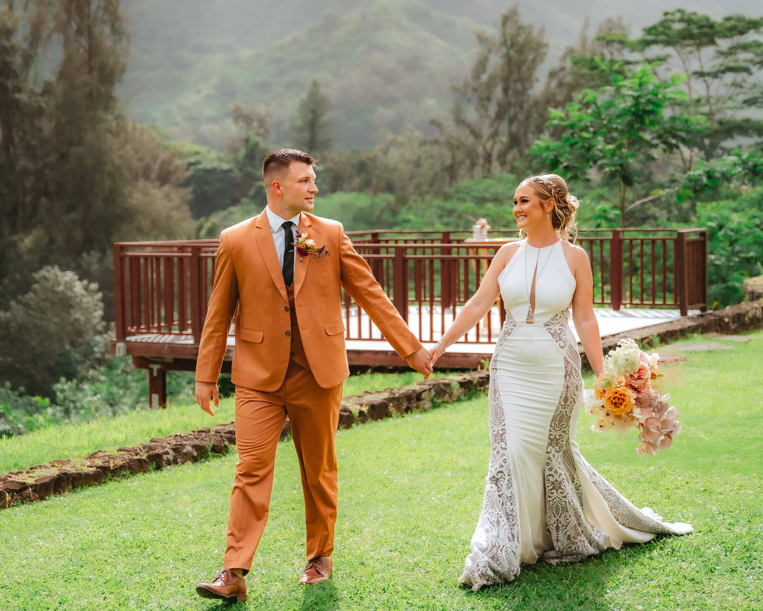 A bride and groom hold hands and walk on a grassy lawn outdoors, with trees and a mountain in the background. The bride wears a white, lace-accented gown and holds a bouquet, while the groom wears a brown suit with a boutonnière.