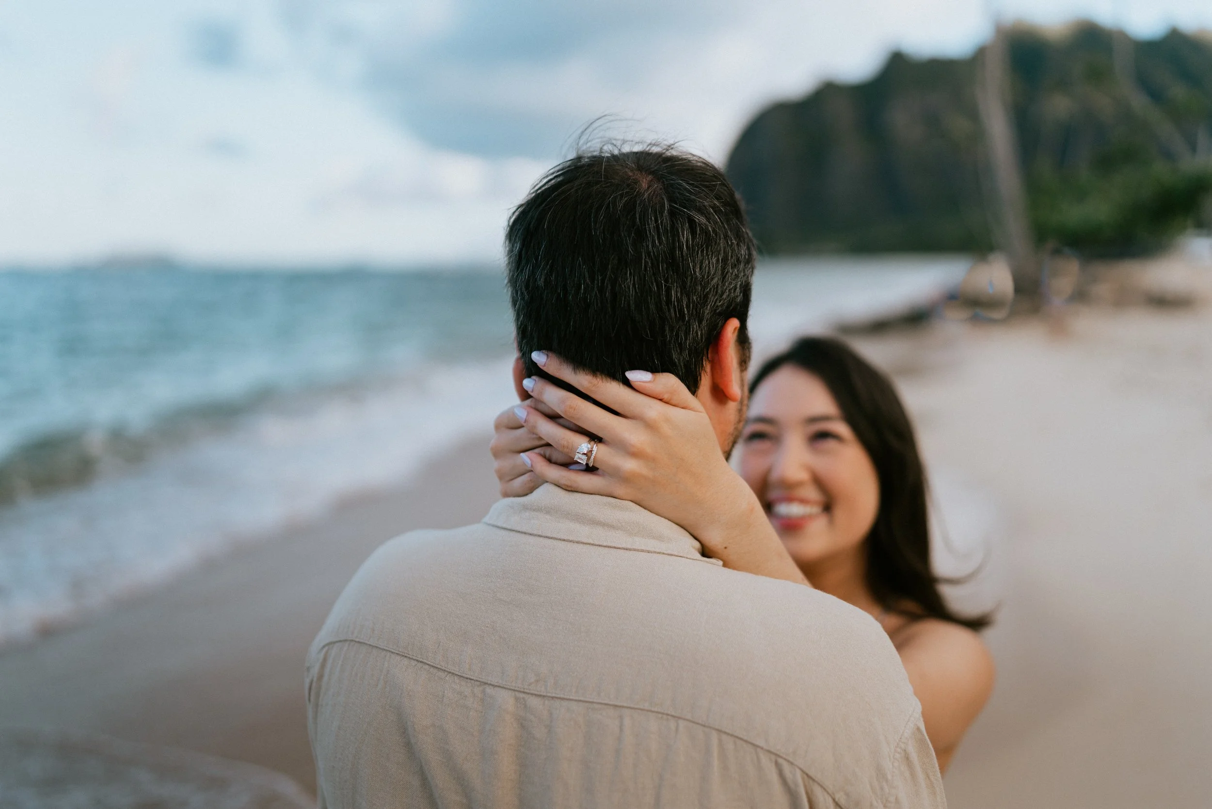 A couple on a beach, with the woman holding the man's face and smiling at him. The man has dark hair and is wearing a light-colored shirt, while the woman has long dark hair and is smiling.
