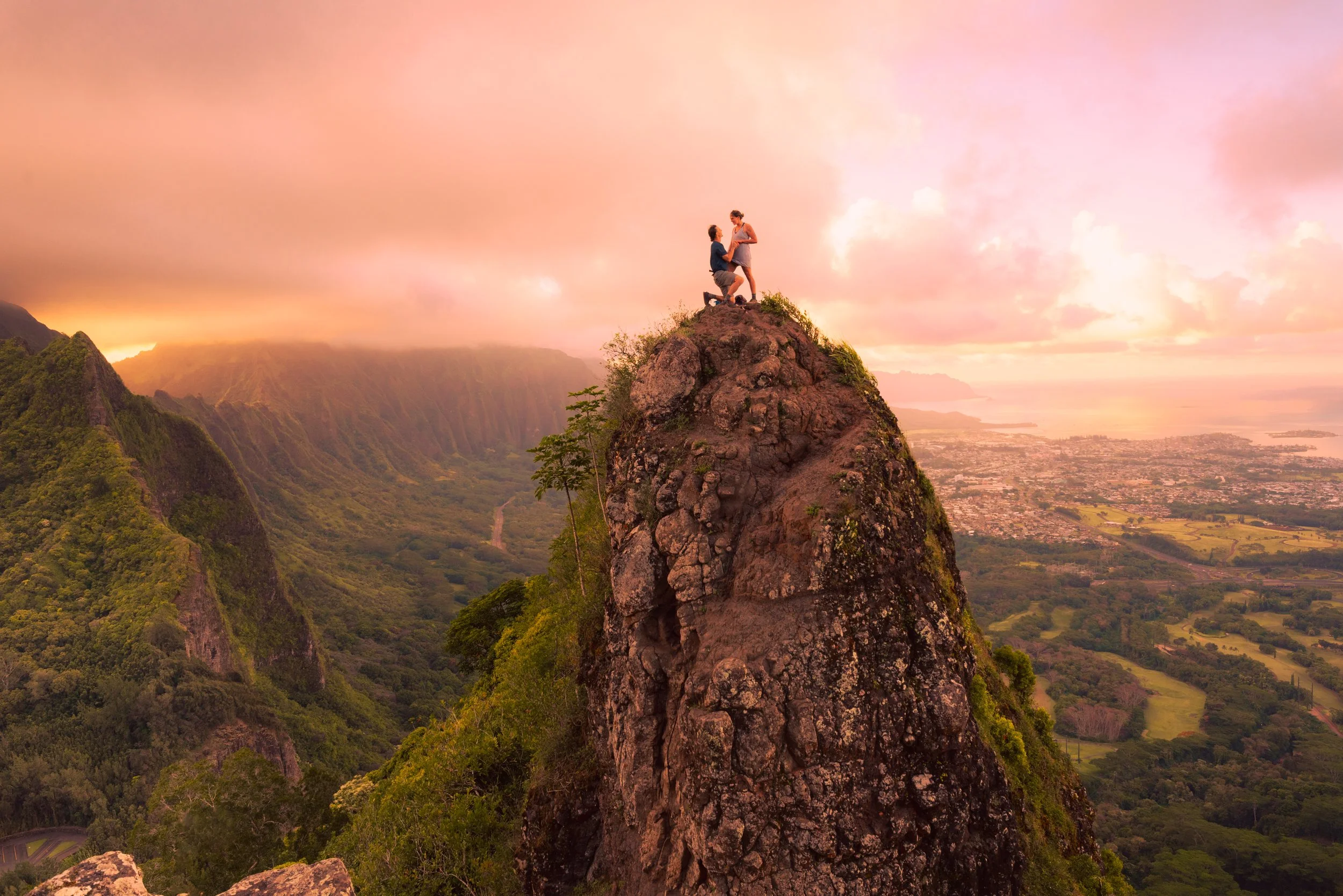 A couple is standing on top of a large rocky peak during sunset or sunrise, with mountains and a valley in the background.