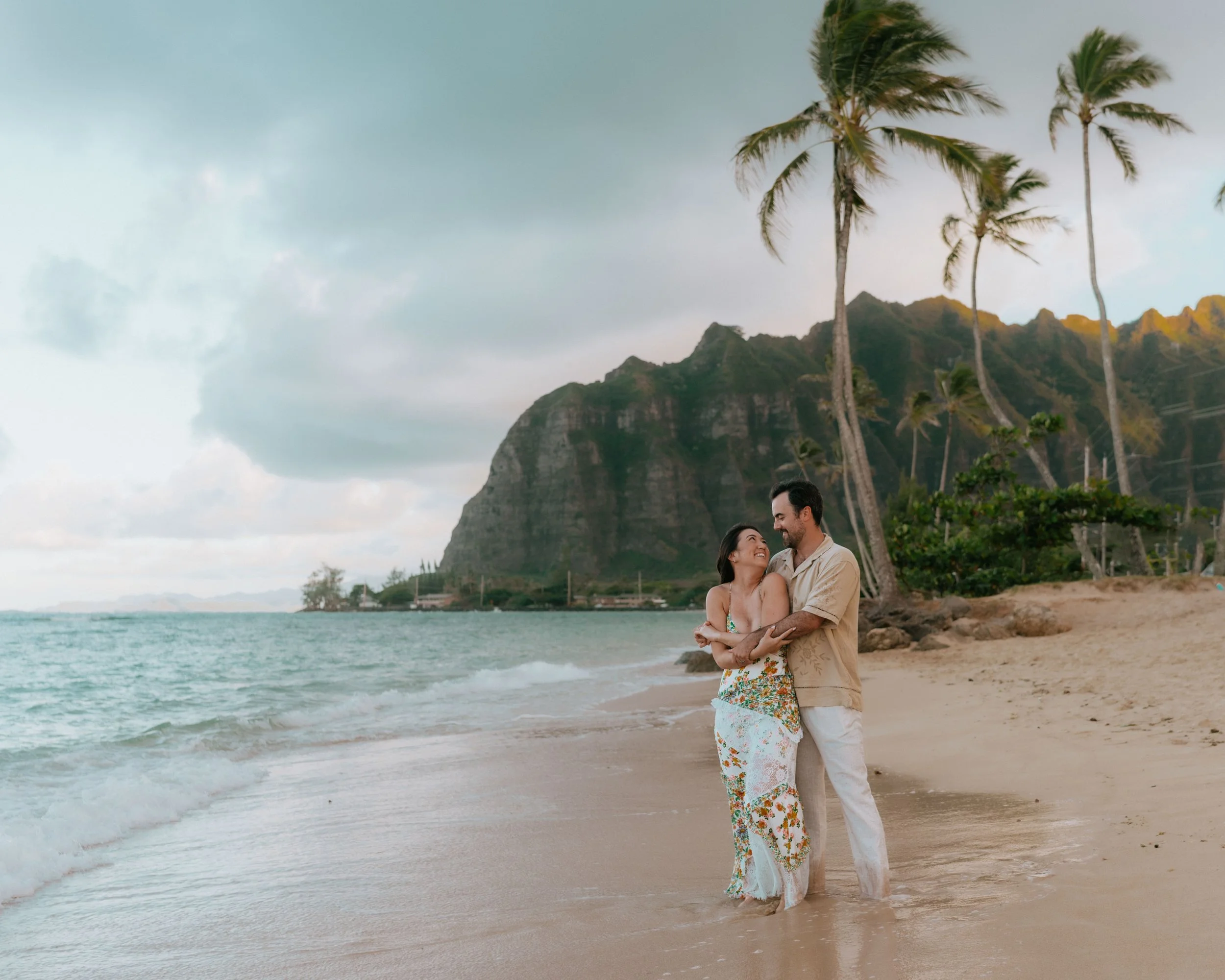 A couple stands embracing on a sandy beach with palm trees and mountains in the background, under a cloudy sky.