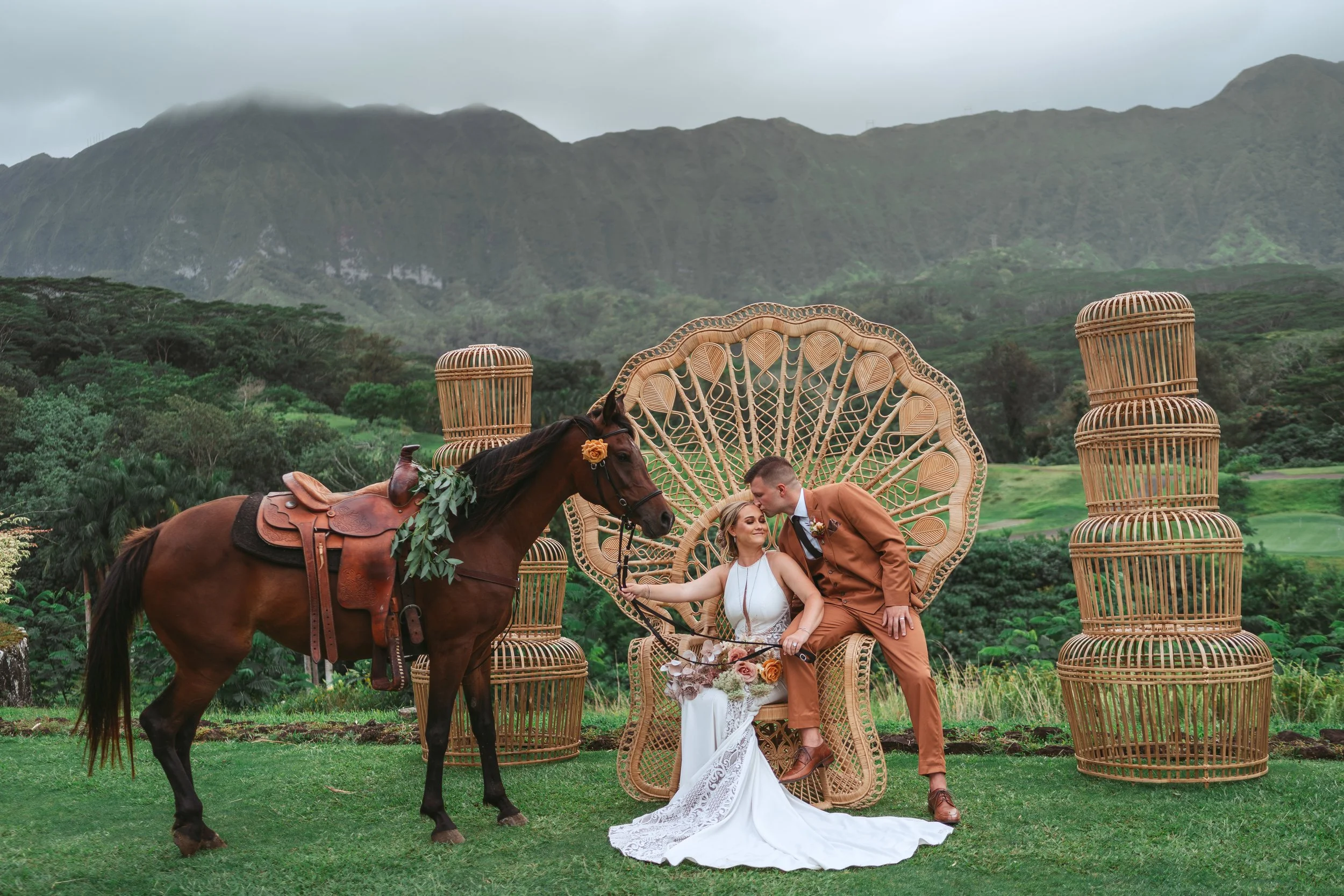 A bride and groom sitting on a wicker chair outdoors, with a horse decorated with greenery and a saddle nearby, and wicker stools in the background, set against green hills and cloudy sky.