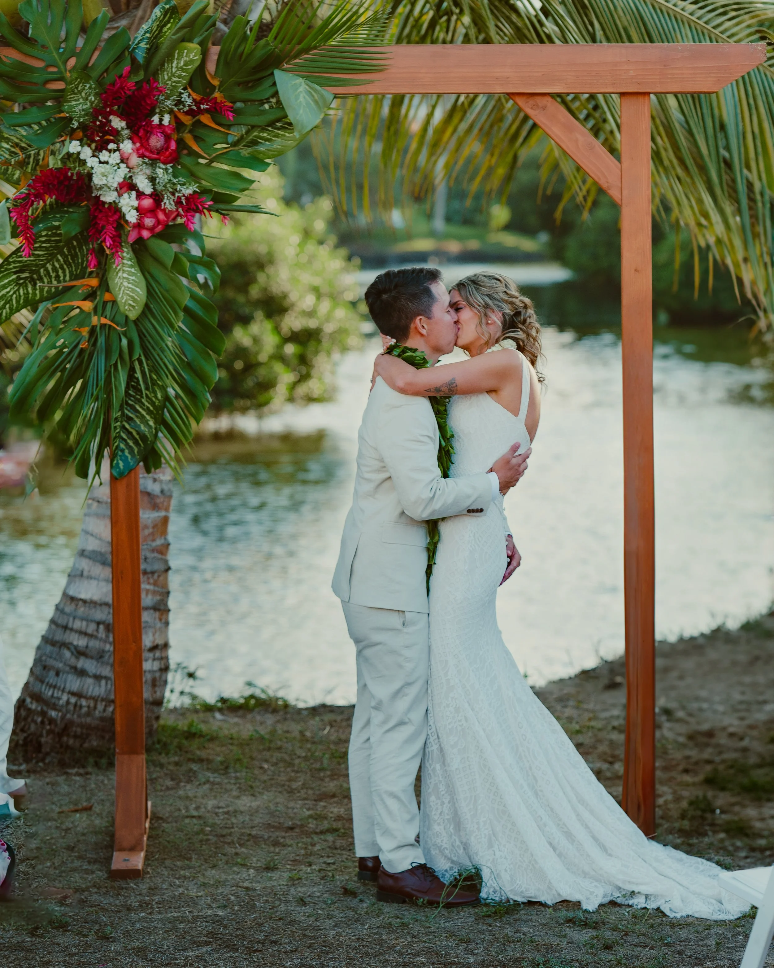 A couple in wedding attire kissing under a wooden arch decorated with tropical flowers and large green leaves near a body of water, with palm trees in the background.