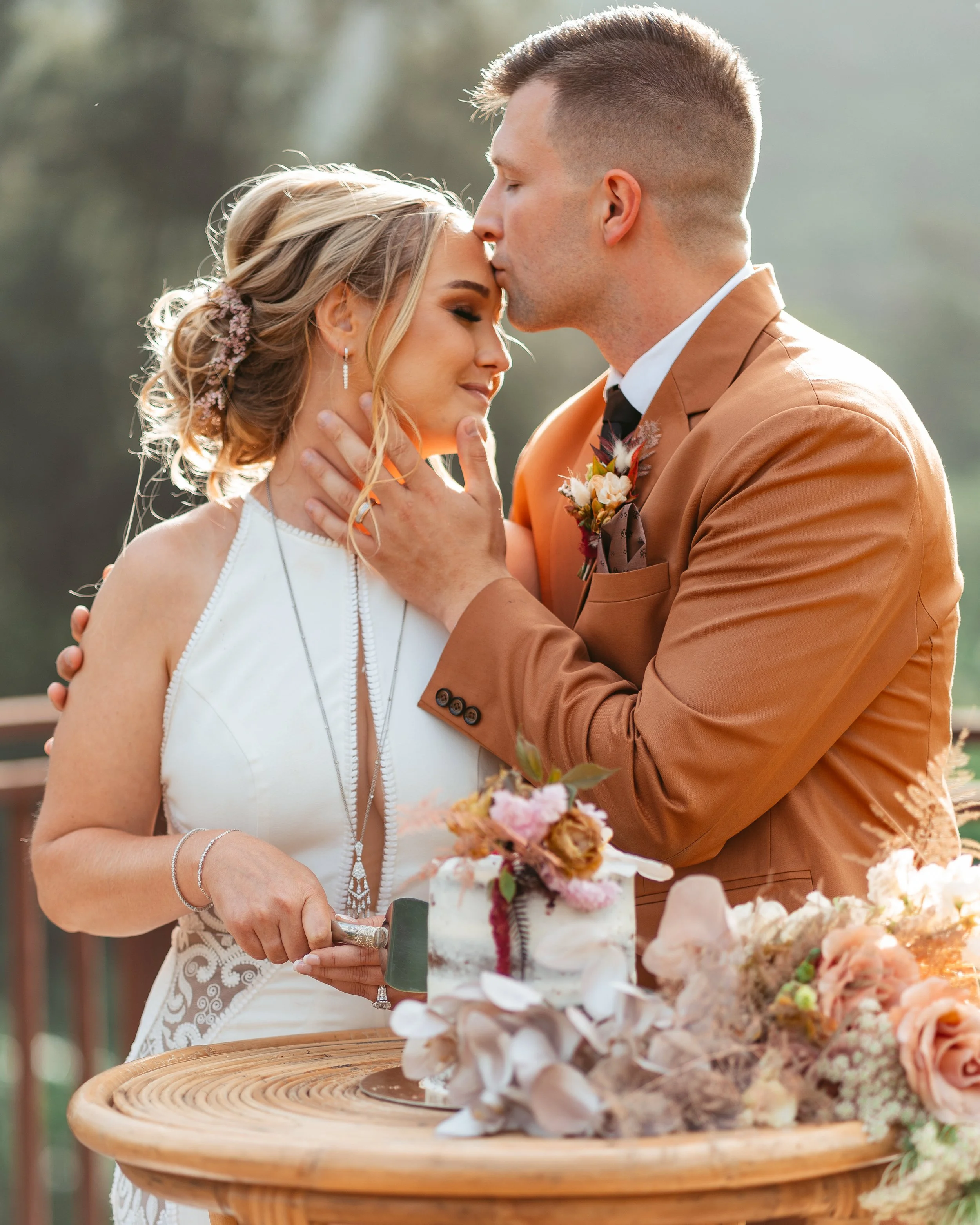 A bride and groom at their wedding, with the groom gently kissing the bride's forehead as she smiles with her eyes closed, holding a cake knife over a decorated wedding cake on a wooden table surrounded by flowers.