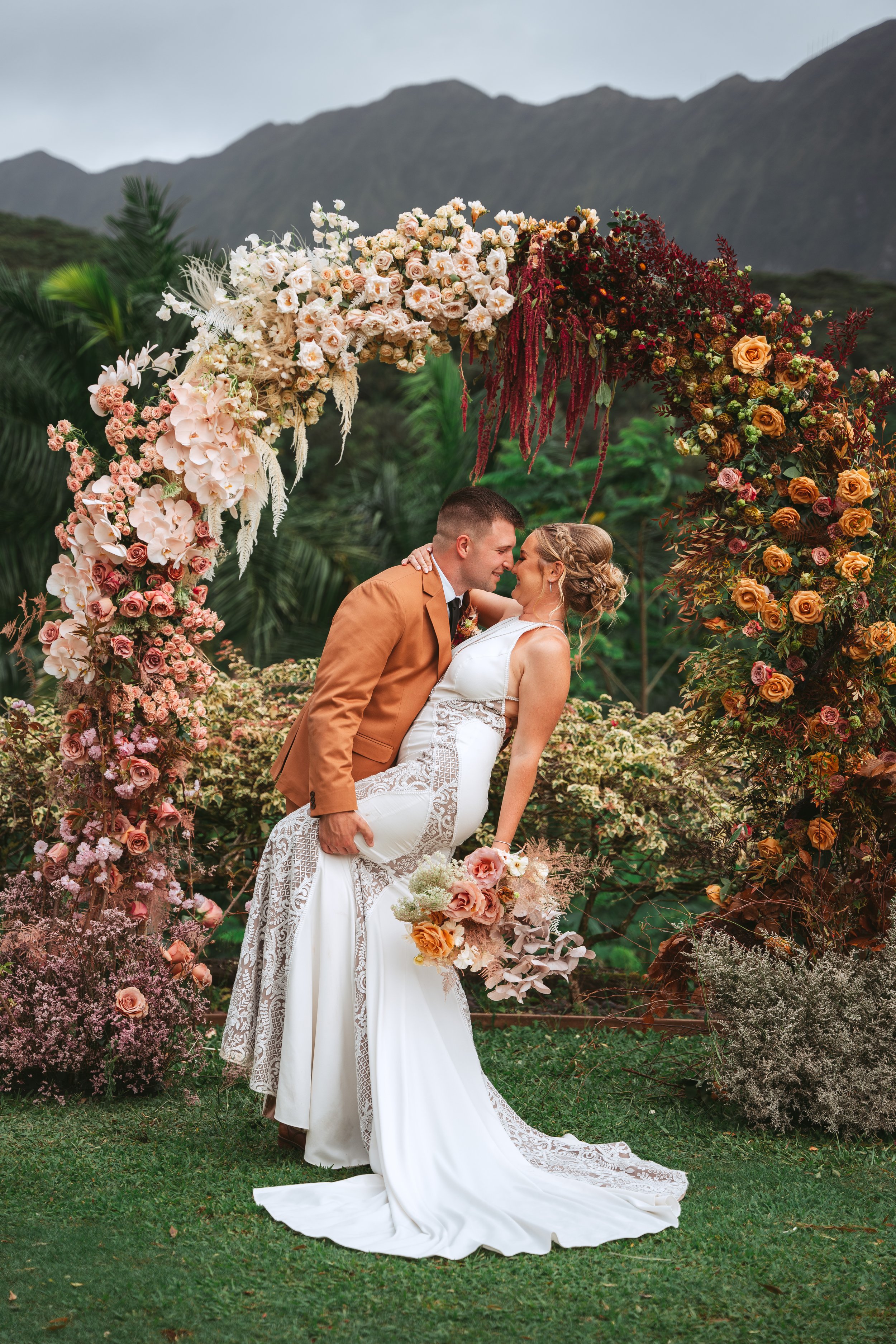 A bride and groom posing under a large arch made of colorful flowers, with lush green landscape and mountain in the background.