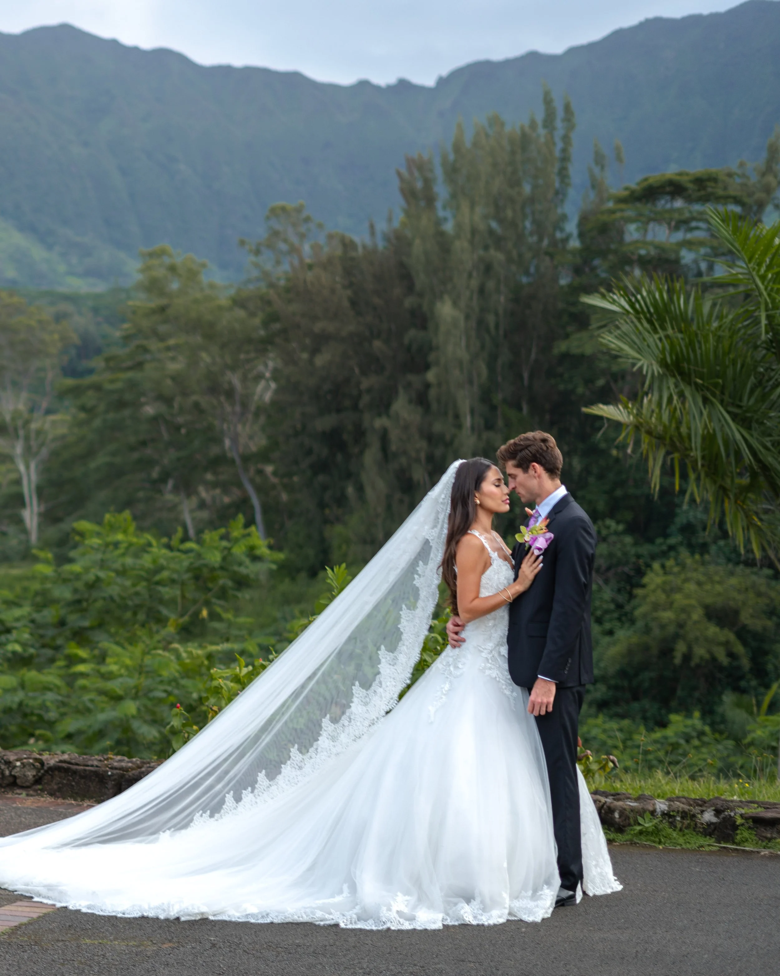 A bride and groom embrace outdoors with lush green mountains and trees in the background, the bride wearing a long white wedding gown and veil, and the groom in a dark suit holding a small bouquet.