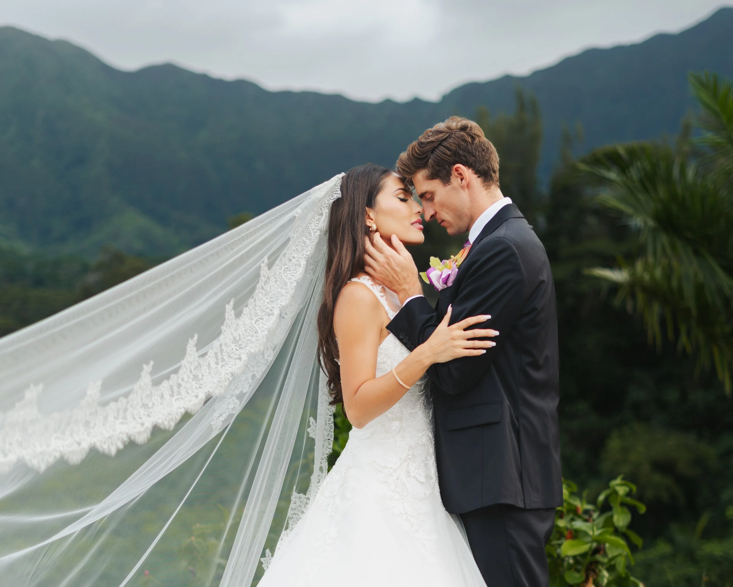 Bride and groom embrace outdoors, mountains and lush greenery in the background, wedding attire, romantic moment.