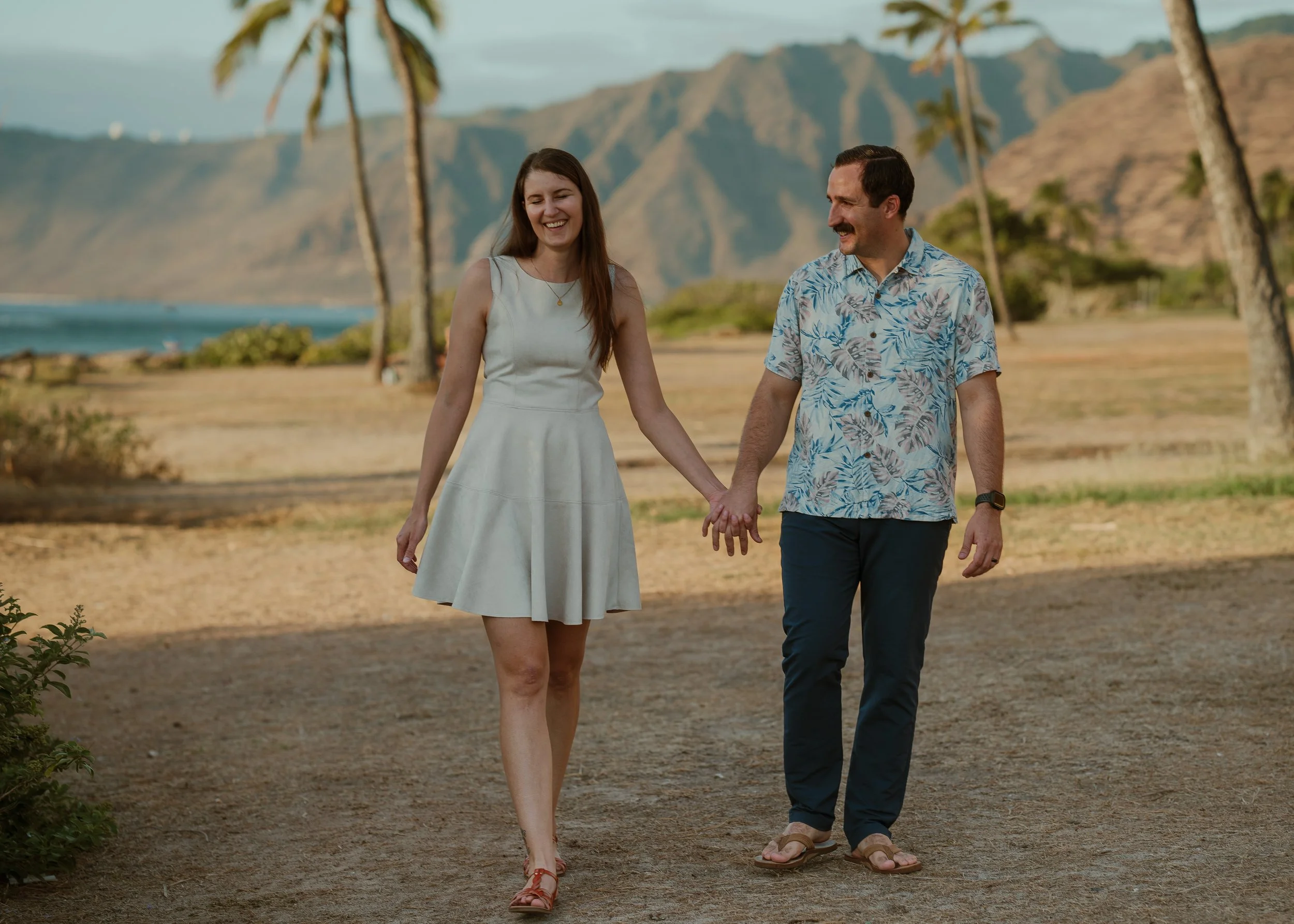 A smiling couple holding hands, walking on a sandy path near the beach with palm trees and mountains in the background.