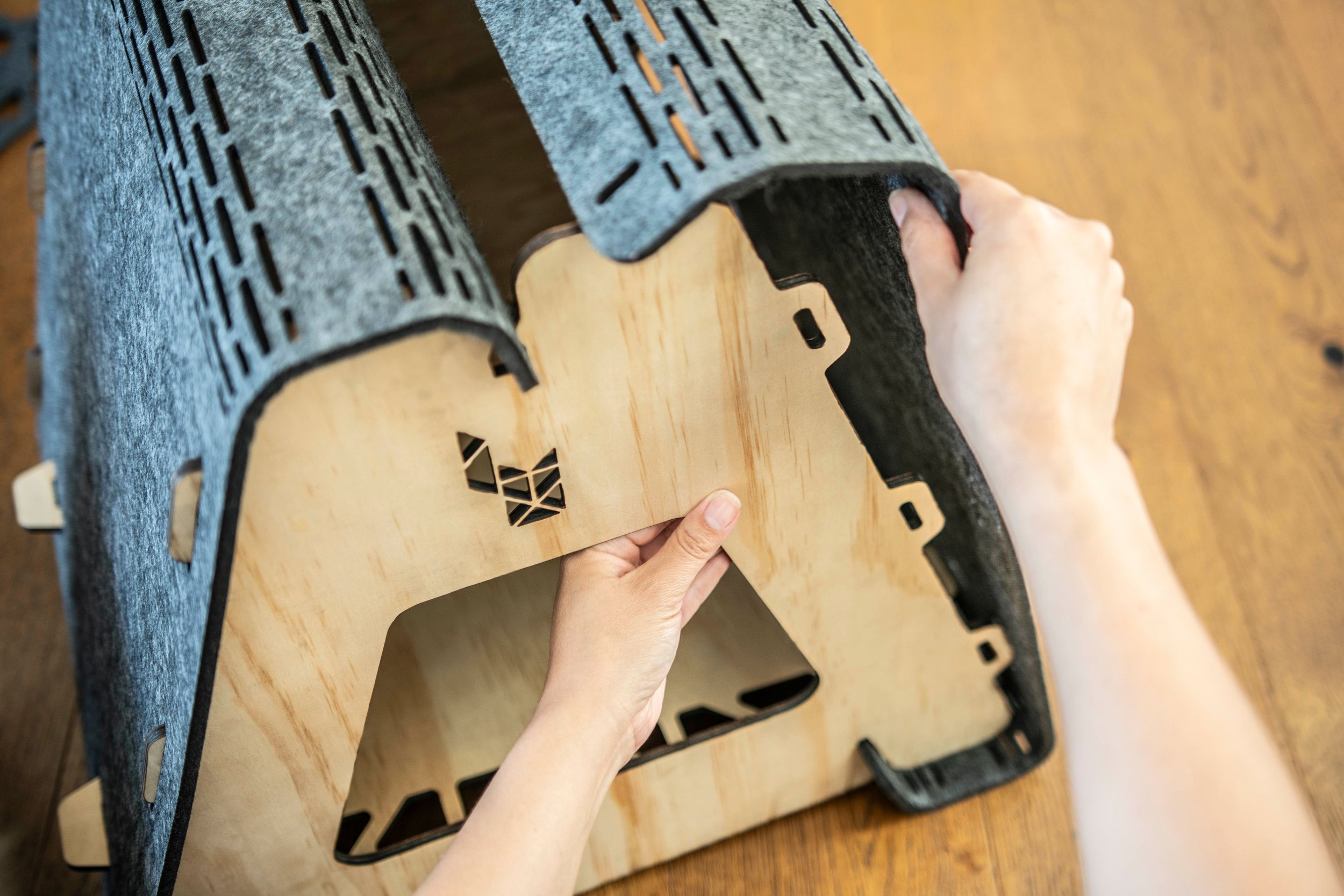 Person assembling a wooden and felt pet house, with hands adjusting the structure on a wooden surface.