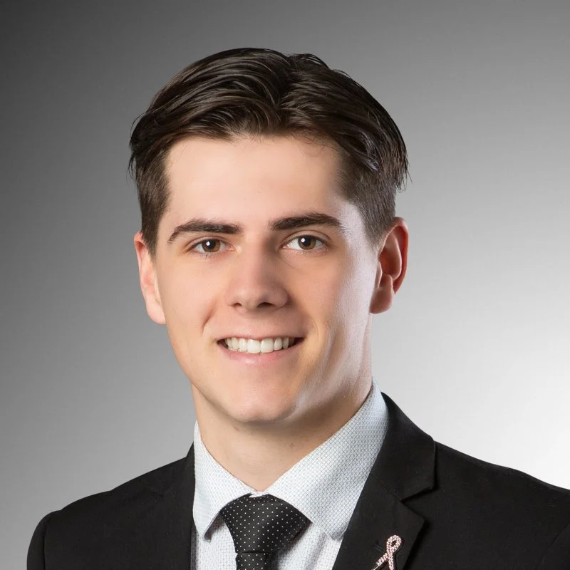 Professional portrait of a young man with brown hair, wearing a black suit, white shirt, and black polka dot tie, smiling against a gray background.